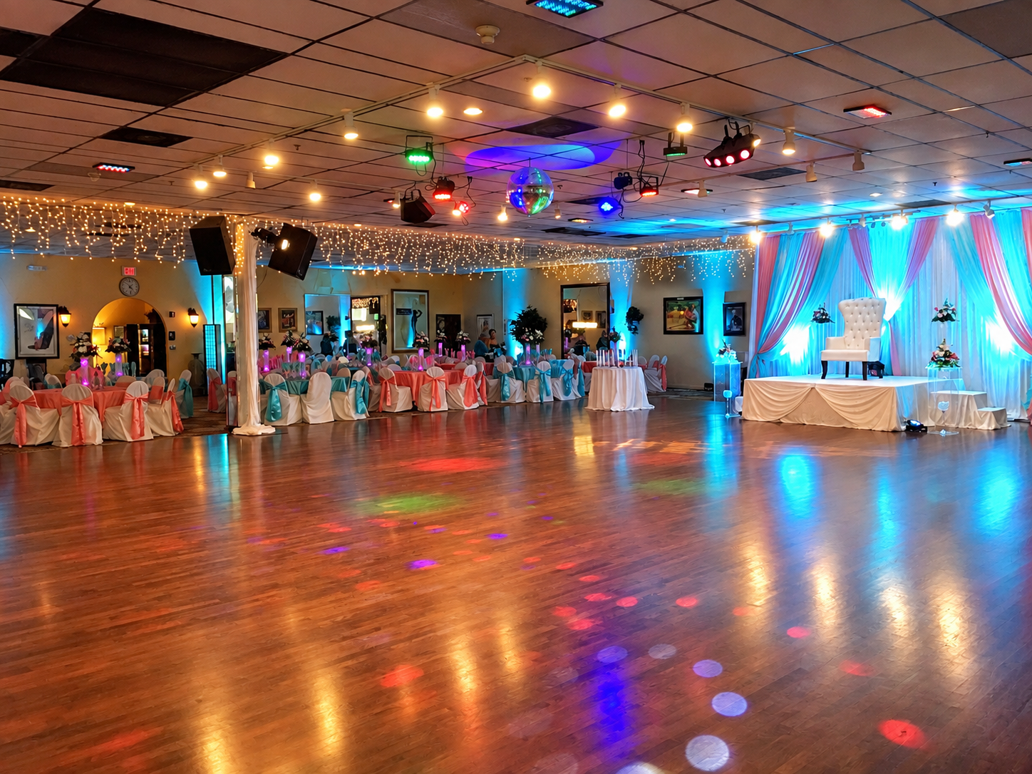 Wedding reception hall with decorated tables and chairs, a stage with a white throne, colorful lights, fairy lights, and a dance floor.