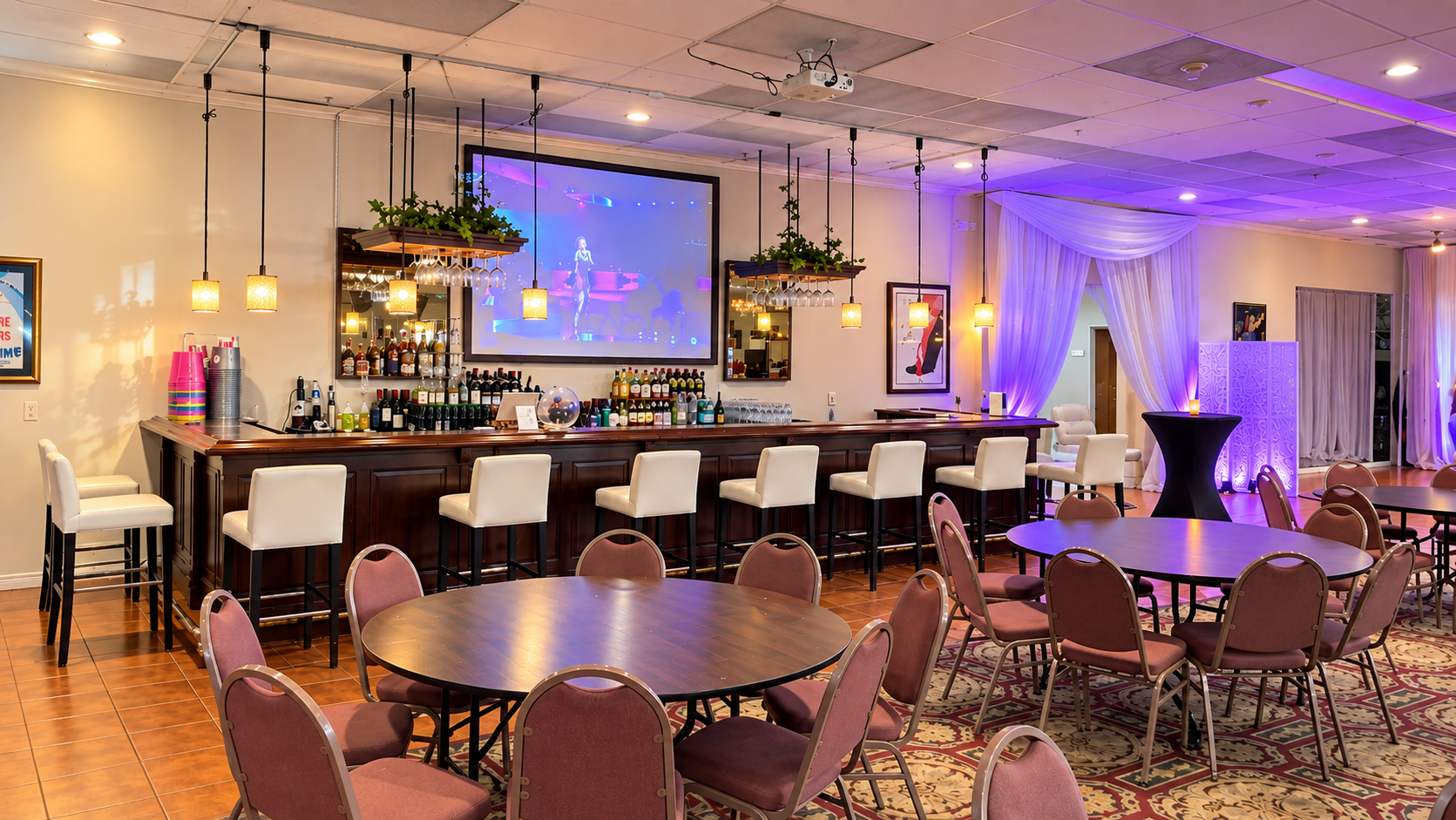 An elegant event space with a bar, round tables, and pink chairs, decorated with purple and white drapery,Artwork, and a large screen displaying a performance.