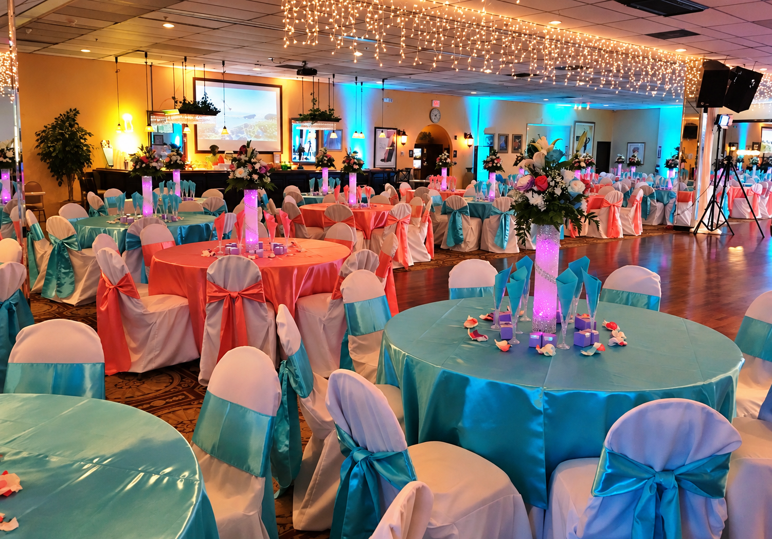 Decorated banquet hall with round tables covered in turquoise and pink satin tablecloths, adorned with large floral centerpieces and colorful sashes on white chairs, under twinkling string lights.