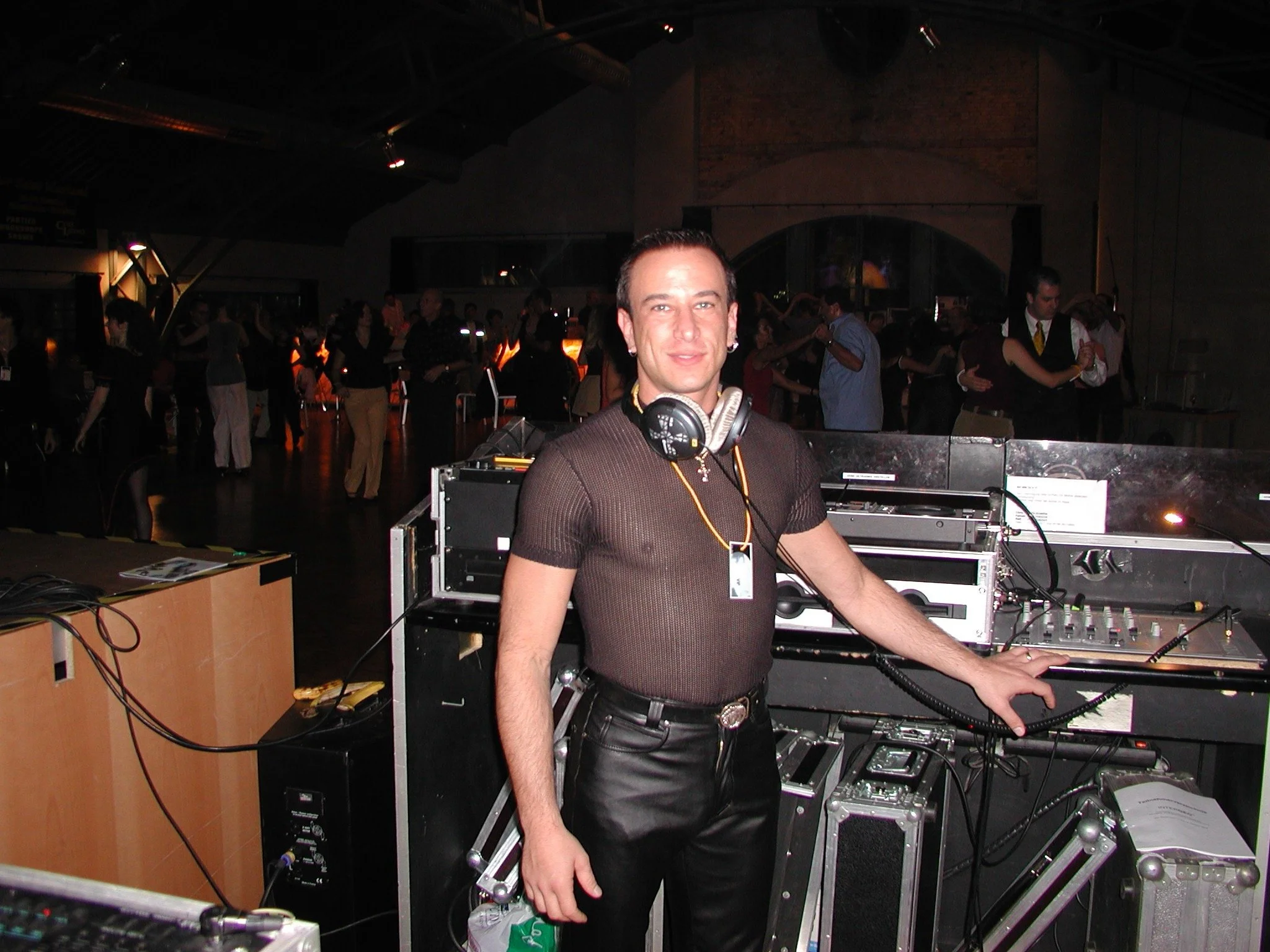 A man with short dark hair, wearing a dark brown shirt and black leather pants, standing at a DJ booth with DJ equipment, surrounded by a crowd dancing at a nightclub or party.