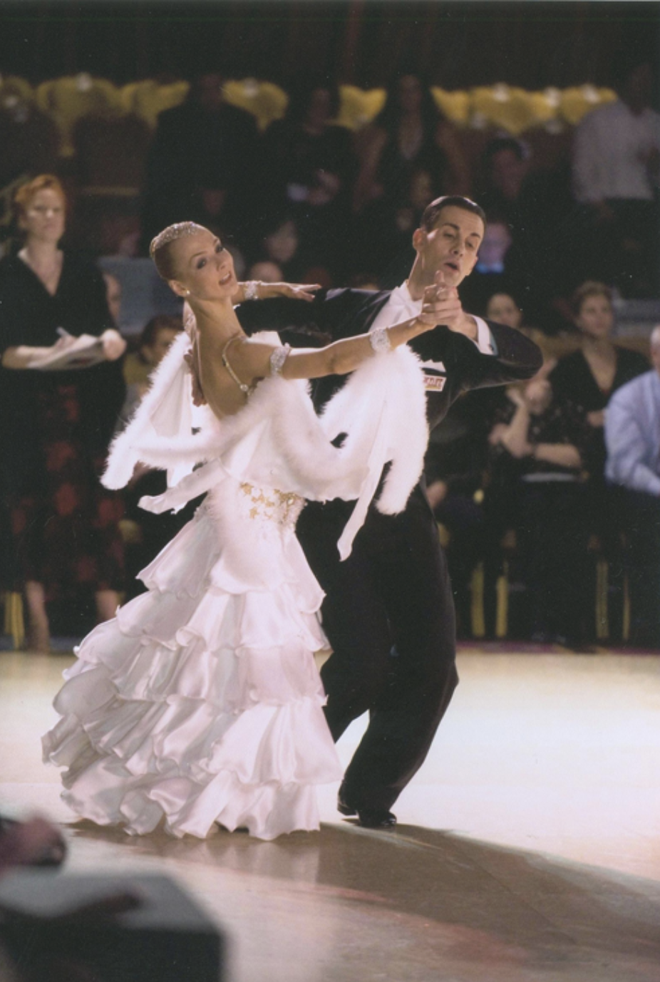 A couple is dancing in formal ballroom attire, with the woman in a flowing white dress and the man in a black suit, during a dance competition.