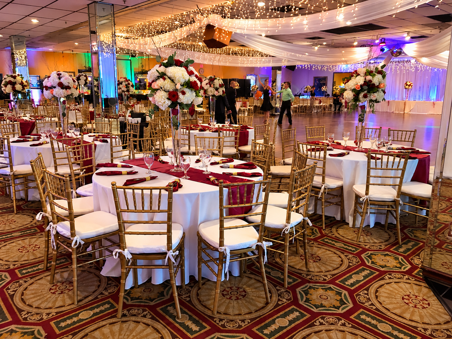 Elegant banquet hall decorated for a celebration with round tables draped in white tablecloths, burgundy table runners, and tall floral centerpieces with white, red, and pink flowers. Gold chiavari chairs with white cushions surround each table. The ceiling is adorned with draped white fabric and string lights, creating a warm, festive atmosphere. In the background, a dance floor and a stage with lighting and sound equipment are visible.