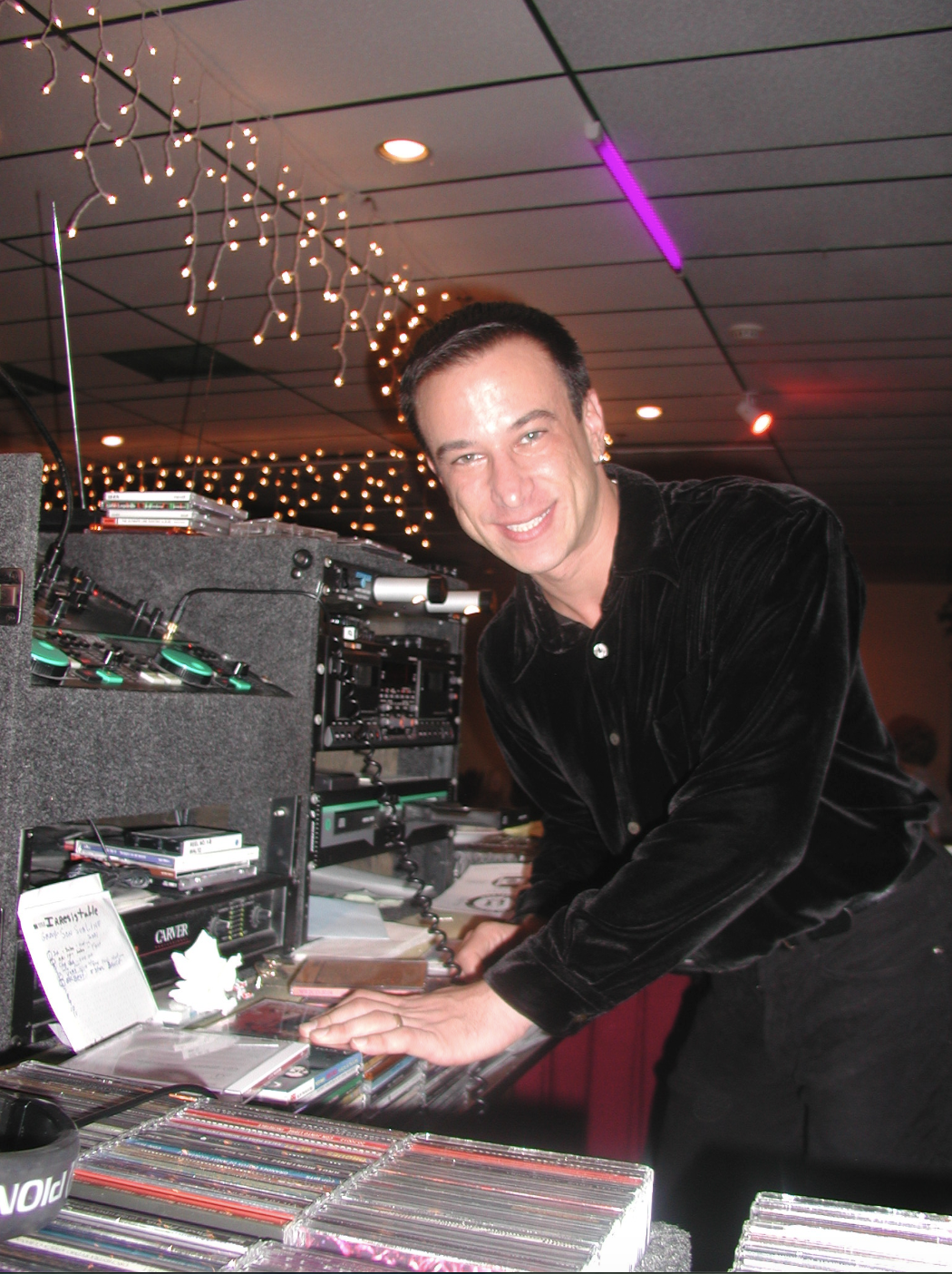 A man in a black shirt smiling at the camera while leaning over a table with DJ equipment and records in a decorated venue with string lights.