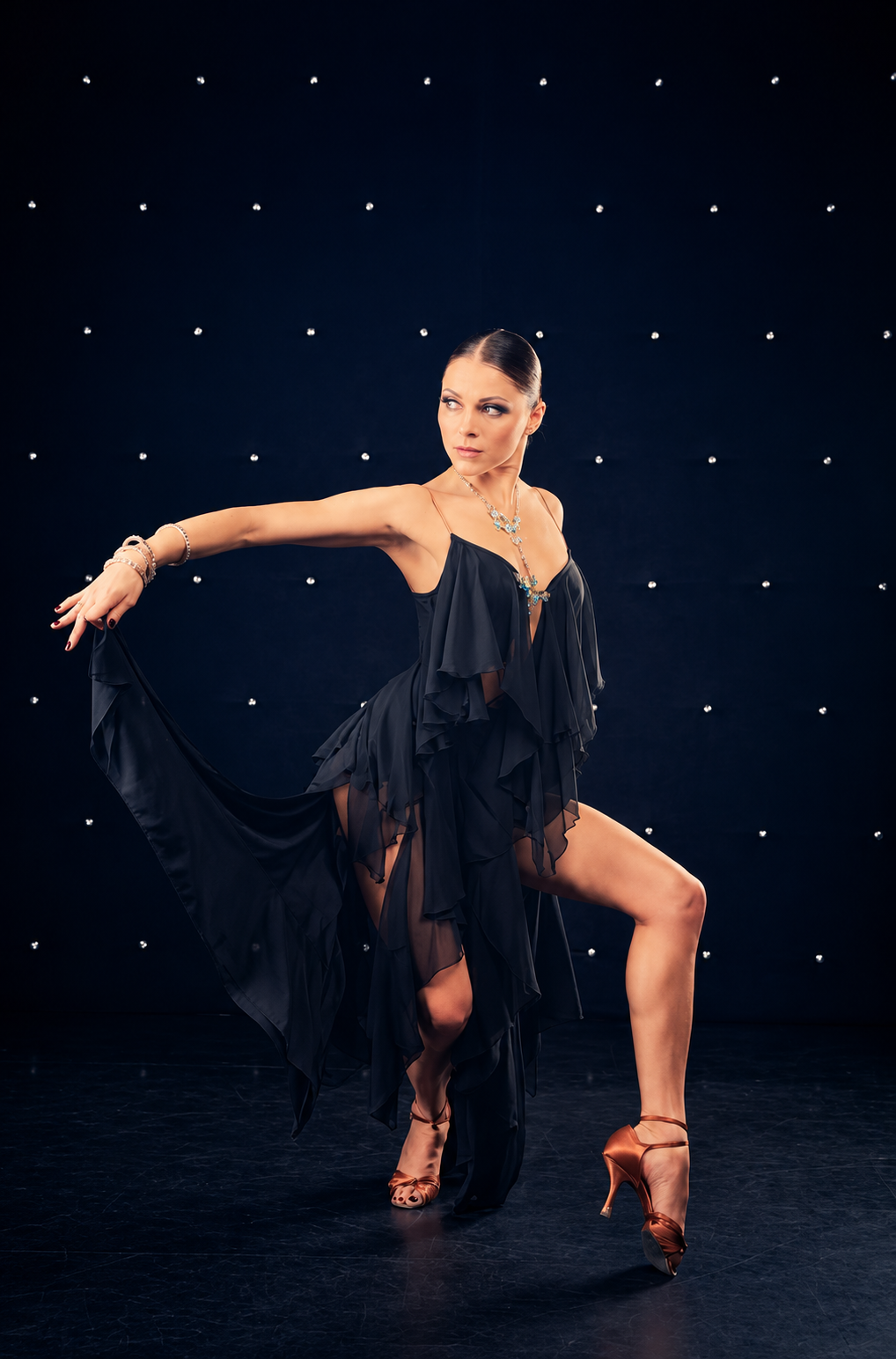 A woman in a black, ruffled dress striking a dance pose on a dark stage with small, illuminated dots in the background, wearing high heels and jewelry.