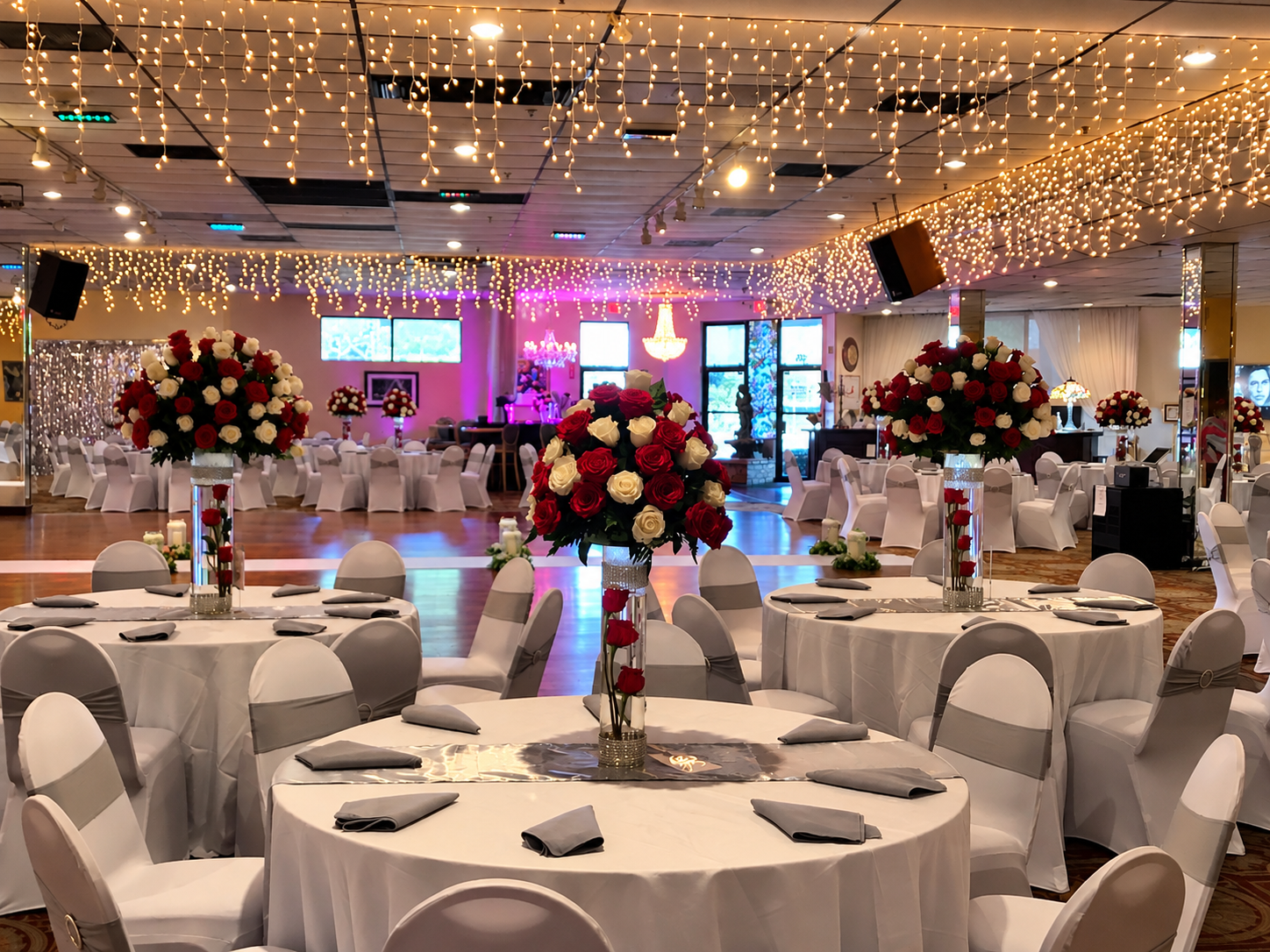 Decorated banquet hall with round tables covered in white tablecloths, each with a large floral centerpiece of red and white roses in a tall glass vase. Chairs are white with gray sashes. Ceiling is adorned with string lights, and the background features a stage with pink and purple lighting, chandeliers, and windowed walls with decorative elements.