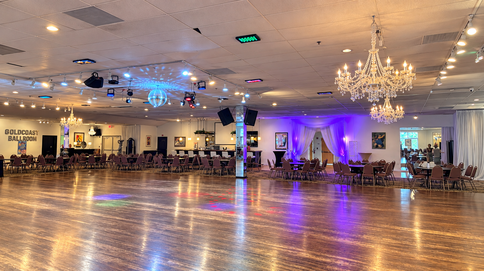 Empty ballroom with many round tables and chairs, chandeliers, purple lighting, a disco ball, and a sign reading 'Gold Coast Ballroom' on the wall.