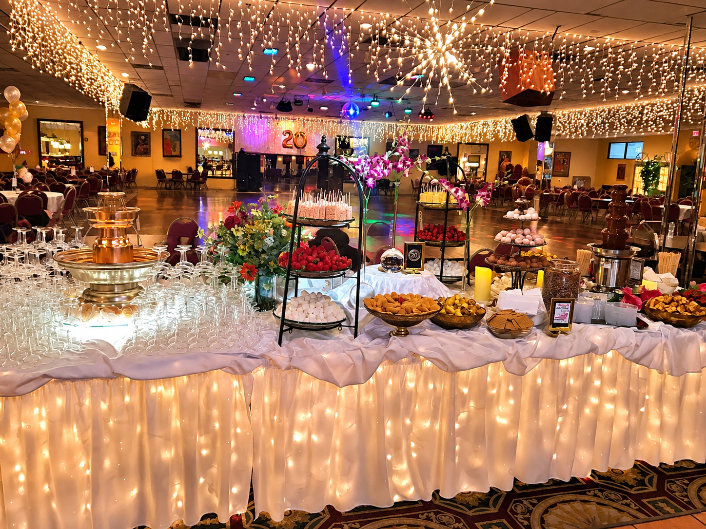 Reception hall decorated with string lights, with a dessert table in the foreground featuring various sweets, flowers, and a fountain for drinks. There are tables with chairs, a dance floor, and balloons in the background, indicating a celebration or party.