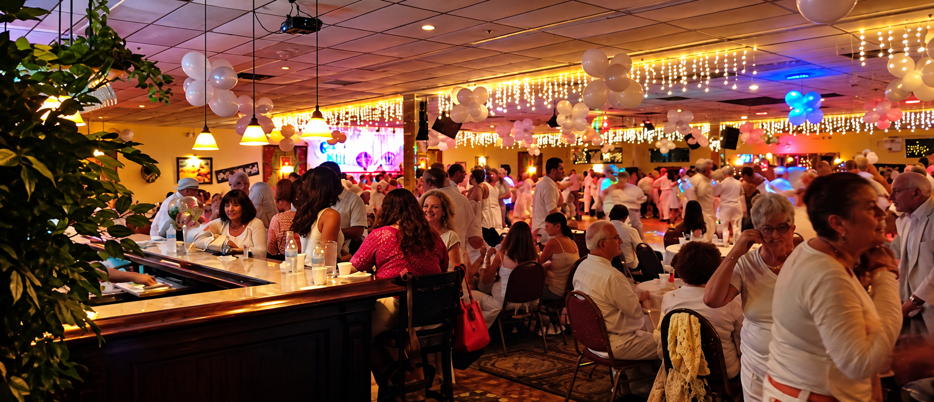 A lively indoor party with people dancing, socializing, and sitting at tables, decorated with balloons and string lights.