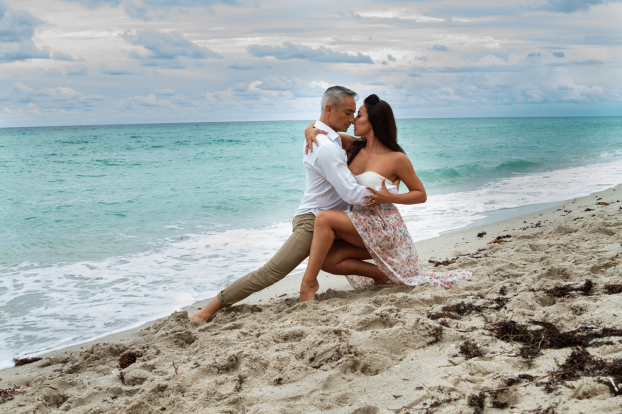 A couple is embracing on a sandy beach with the ocean and cloudy sky in the background.