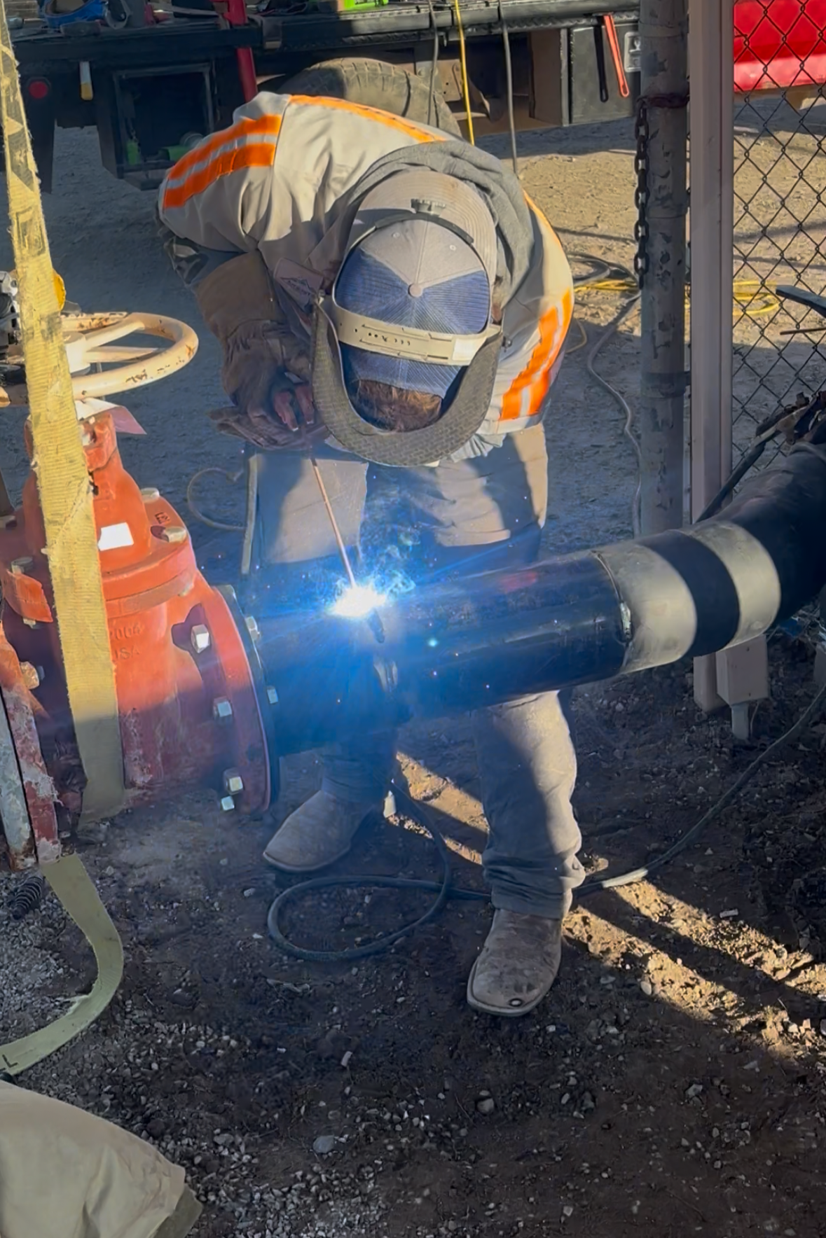 Worker welding a large black pipe at a construction site, wearing protective gear including a welding helmet, gloves, and durable boots.