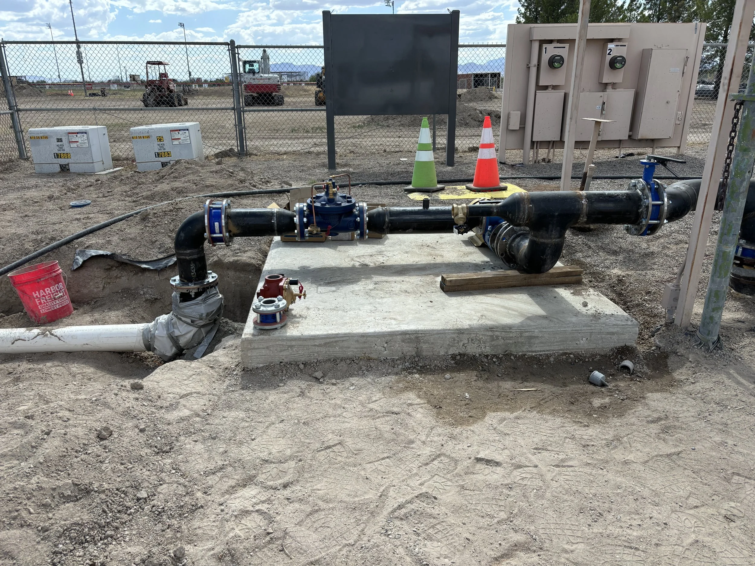 Construction site with exposed pipes and electrical boxes, orange and green safety cones, chain-link fence in the background, and a blue sky with clouds.
