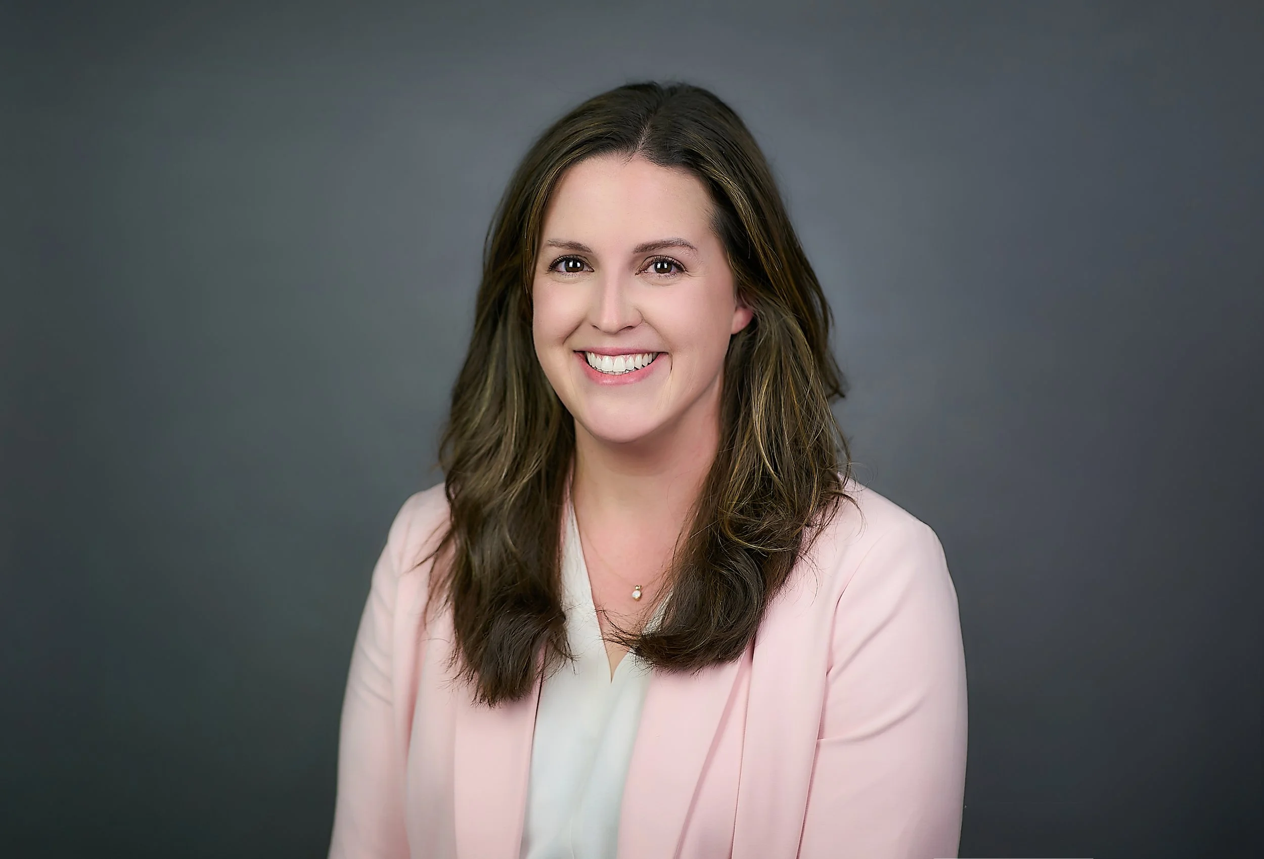 A woman with long brown hair, smiling, wearing a light pink blazer over a white top, against a gray background.