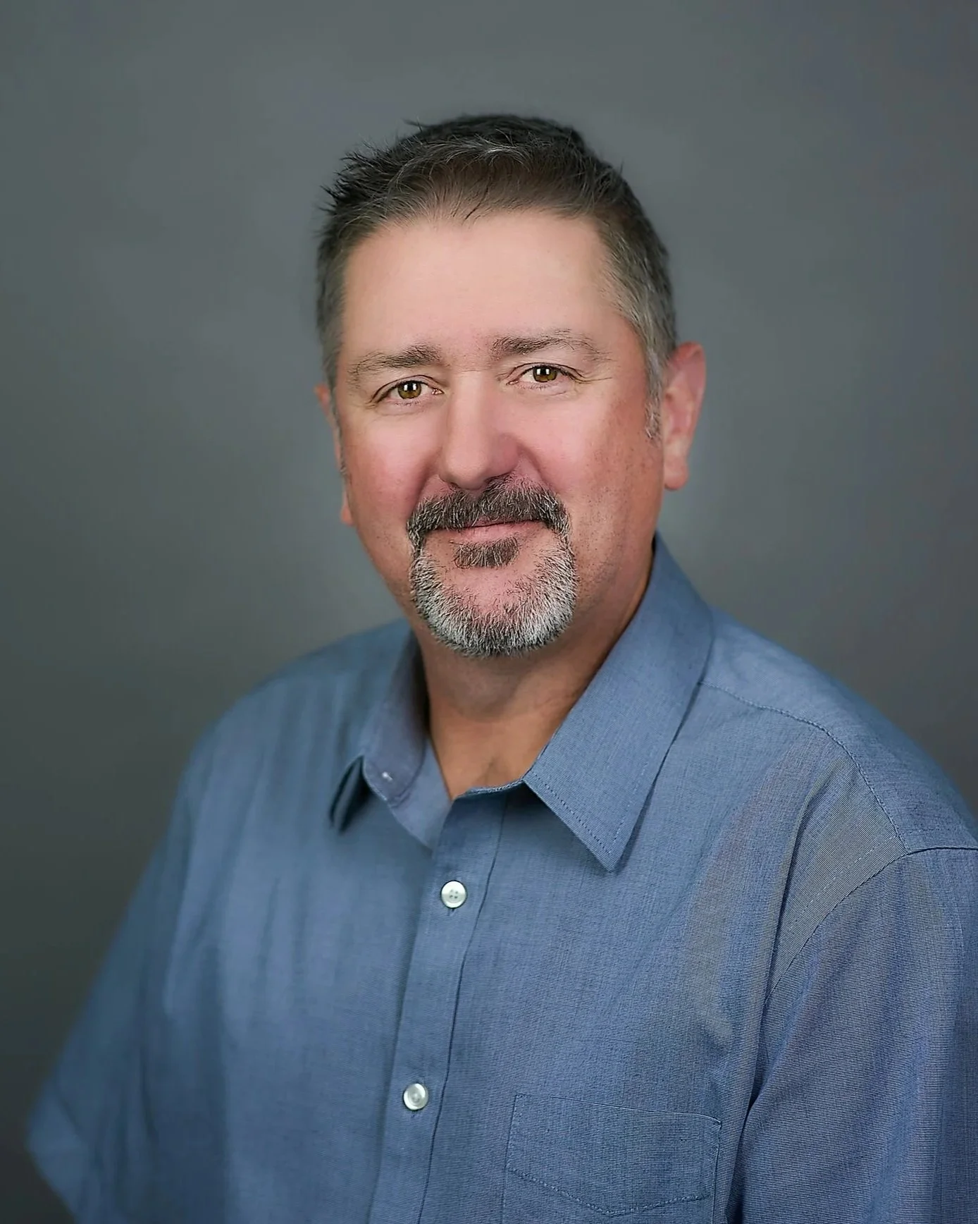 Headshot of a smiling middle-aged man with short brown hair and a beard, wearing a blue button-up shirt against a gray background.