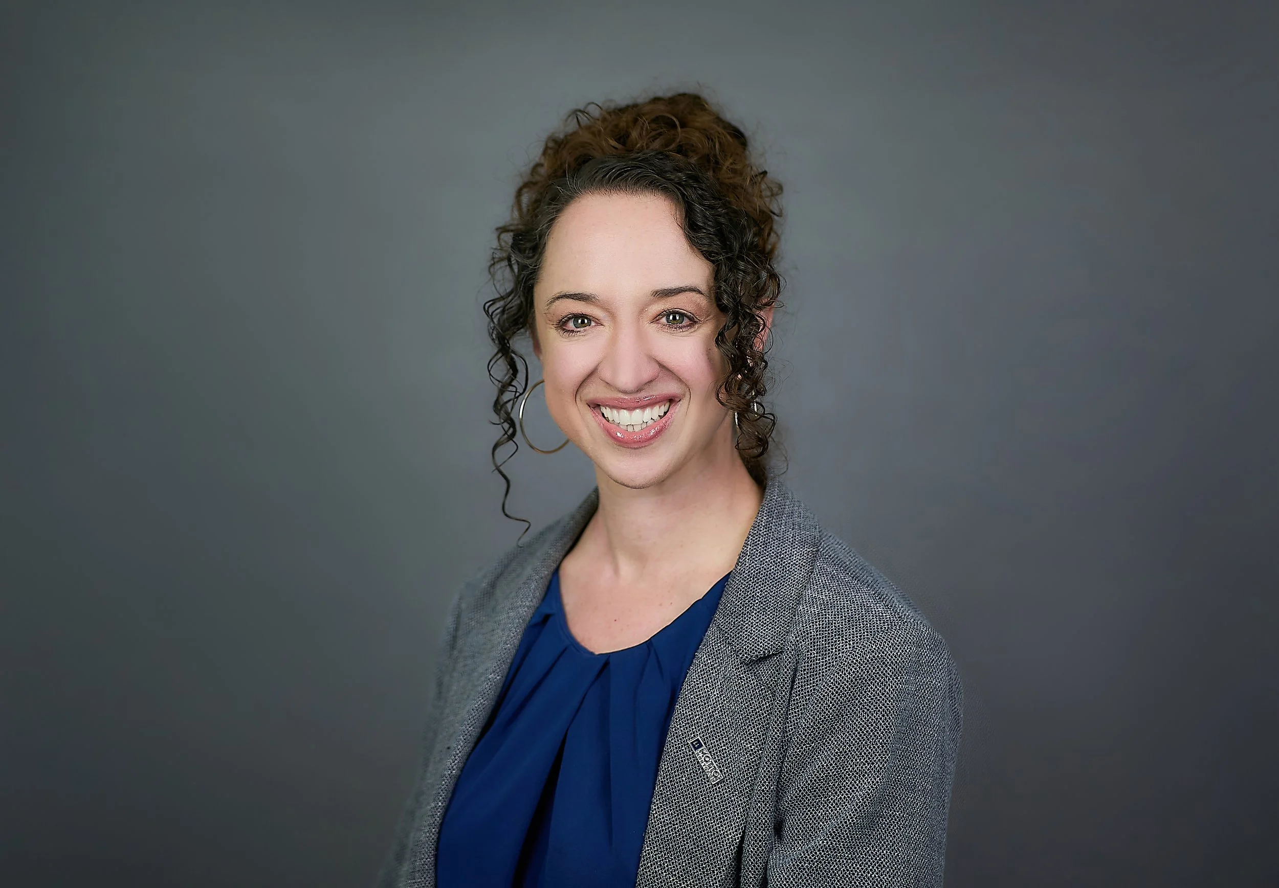 A smiling woman with curly brown hair tied up, wearing a gray blazer over a blue blouse, against a gray background.