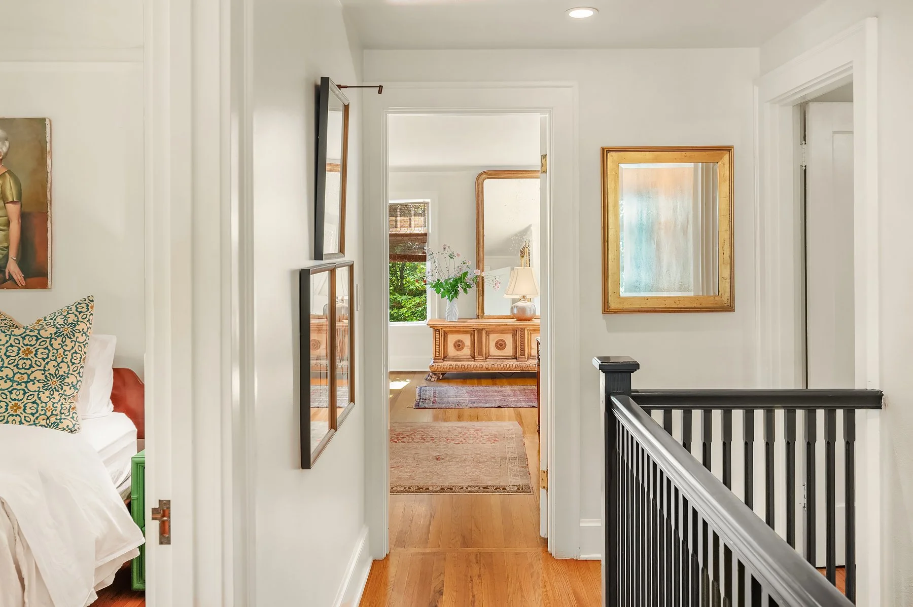 View of a hallway with a staircase, leading to a room with a wooden dresser, a lamp, a vase with flowers, and a window with plants outside.