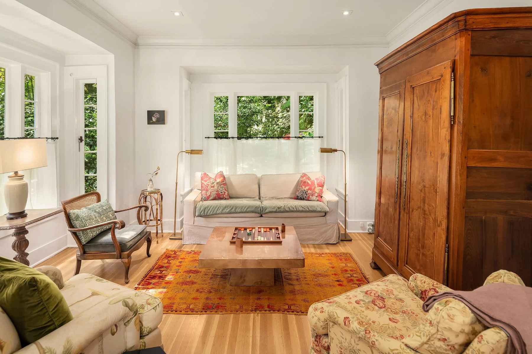 Living room with white walls and wooden furniture, including a large wooden wardrobe on the right. There is a beige sofa with decorative pillows, a wooden coffee table with a backgammon game, a vintage armchair, a small side table with a lamp, and la