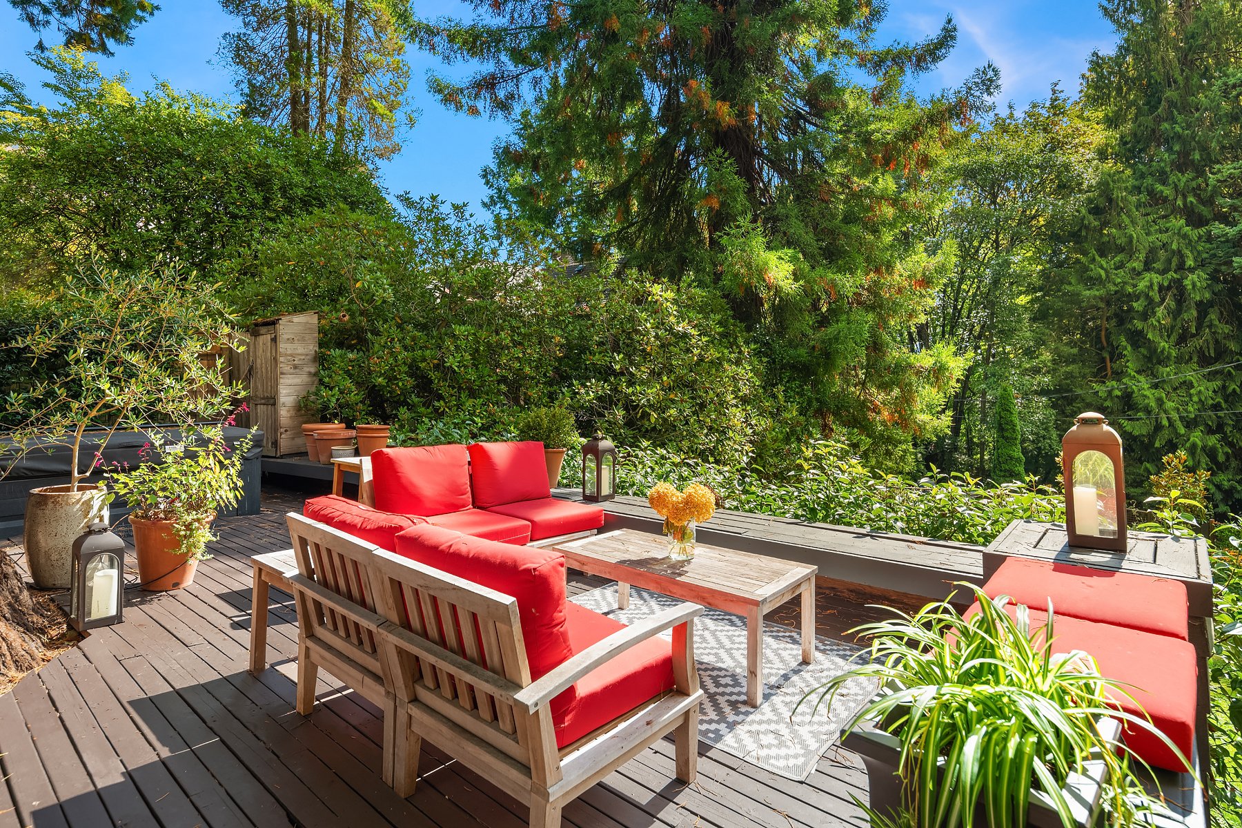 A cozy outdoor deck surrounded by lush green trees and plants with a wooden table, red cushioned chairs, lanterns, and potted plants, set on a clear sunny day.