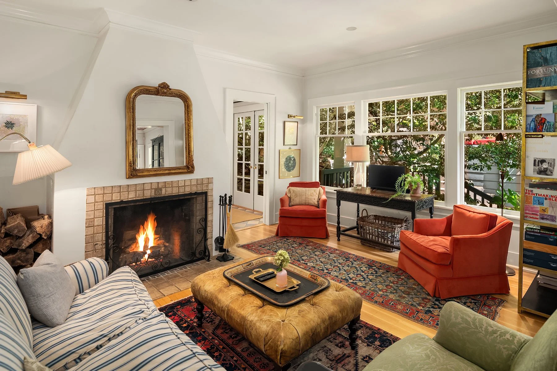 A cozy living room with a lit fireplace, a gold-framed mirror above it, and a wood basket filled with logs to the left. The room features a striped couch with pillows, a tufted ottoman with a tray and remote, and multiple colorful armchairs. Large wi