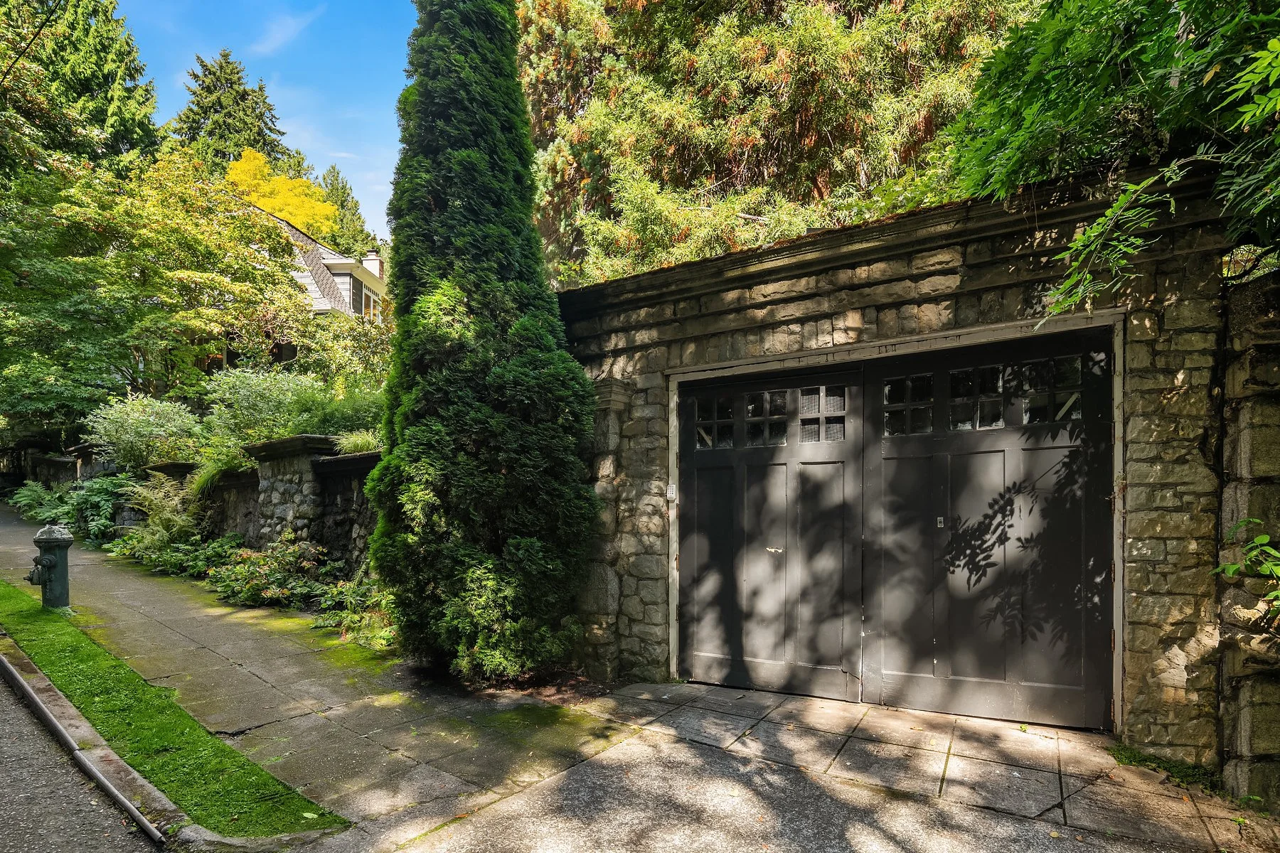 A stone garage with a black door surrounded by lush green trees and shrubs on a sunny day in a residential neighborhood.