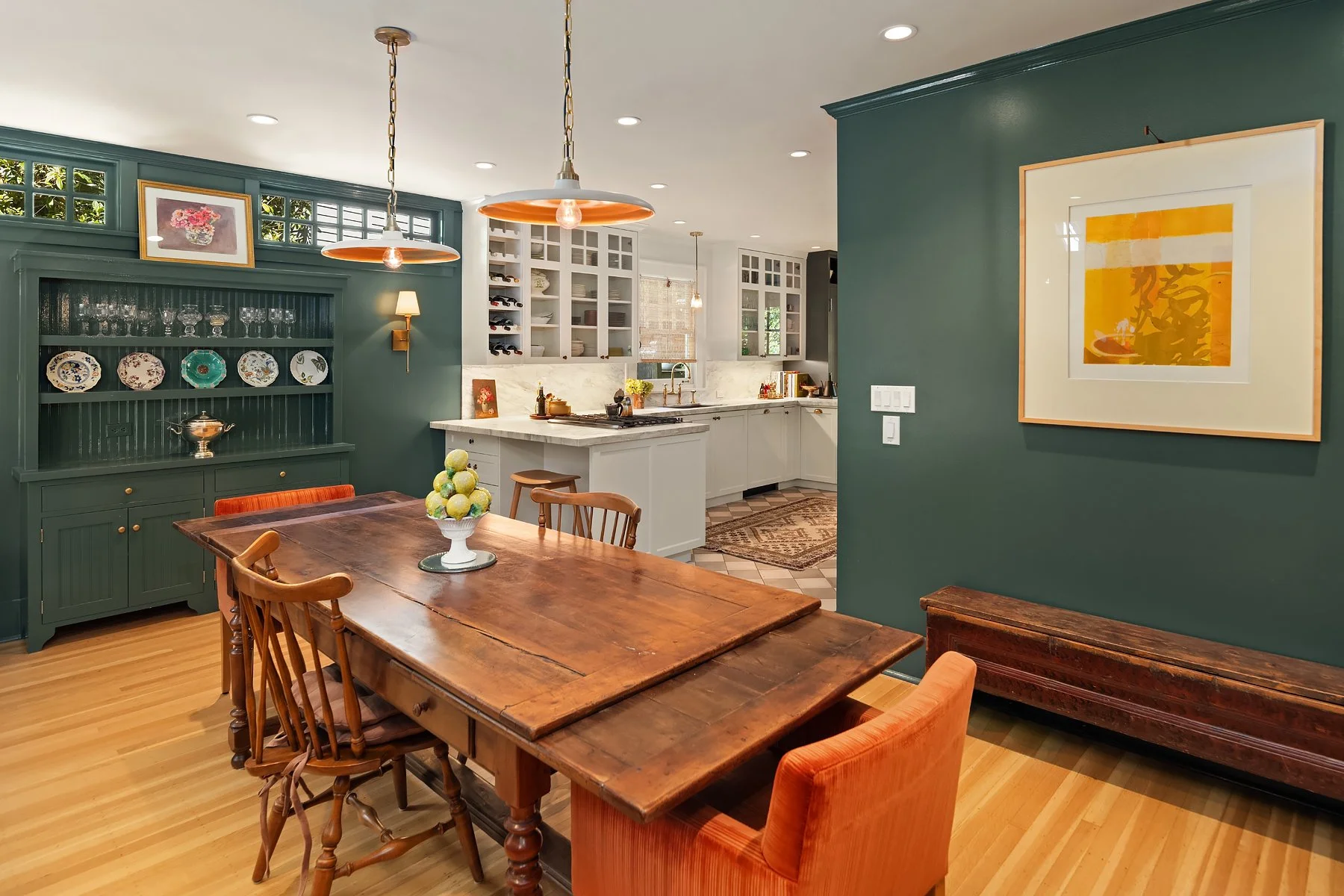 A dining area with a wooden table and six chairs, a green cabinet with decorative plates and glassware, and a kitchen with white cabinets, open shelving, and a marble countertop. The room has hardwood floors, hanging pendant lights, framed artwork on