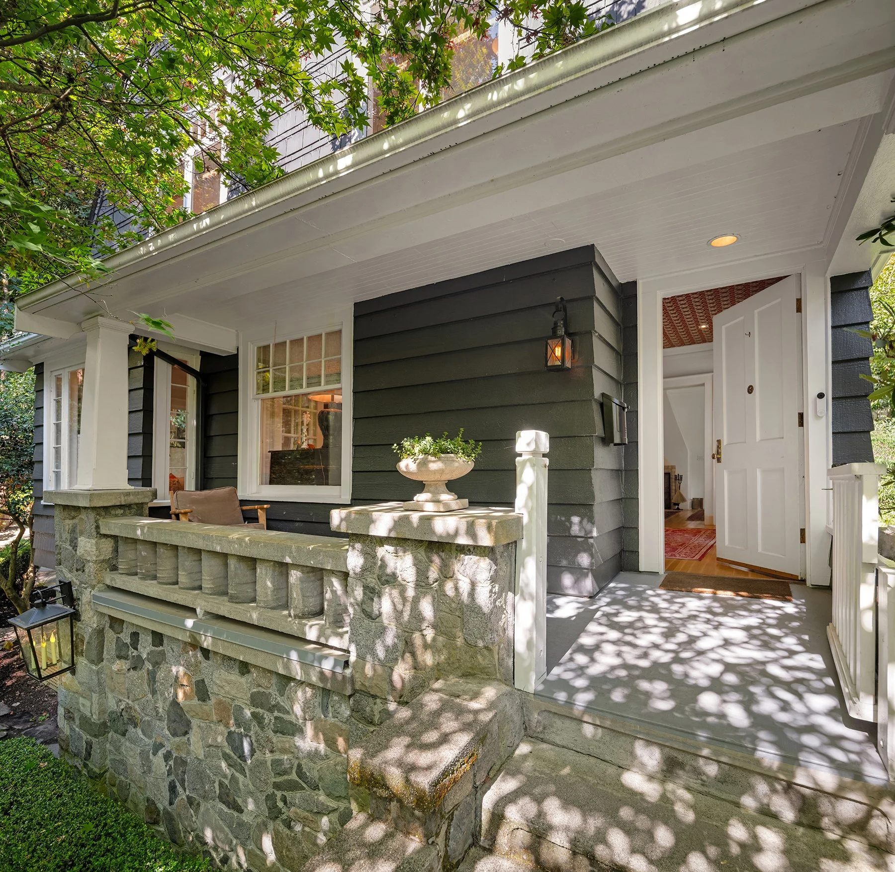 Front porch of a house with dark gray siding, stone foundation, white trim, and an open door revealing a cozy interior with red patterned rug. Potted plants and a lantern-style light fixture are present, surrounded by lush green trees casting shadows