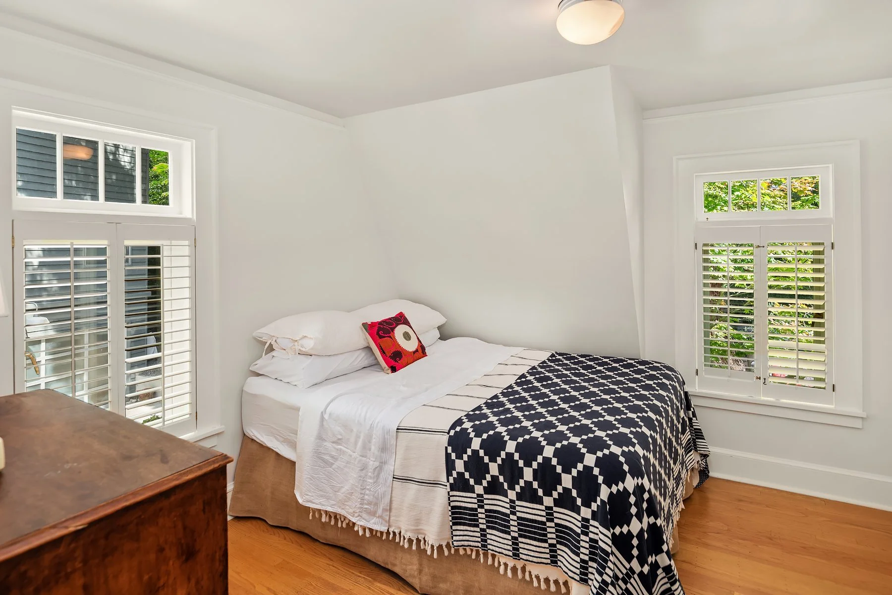 A bedroom with white walls, two large windows with white shutters, and a bed with white bedding and a black-and-white patterned blanket. There is a colorful pillow on the bed, and a wooden dresser is partially visible in the foreground.