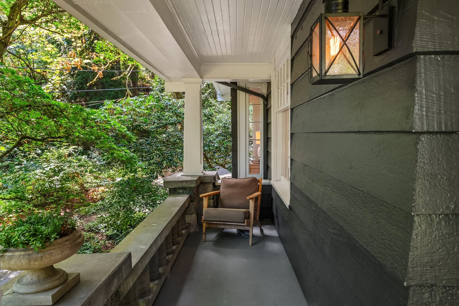 A cozy front porch with a dark painted wooden exterior wall, a wall-mounted lantern, a wooden chair with a cushion, and potted plants with green foliage.