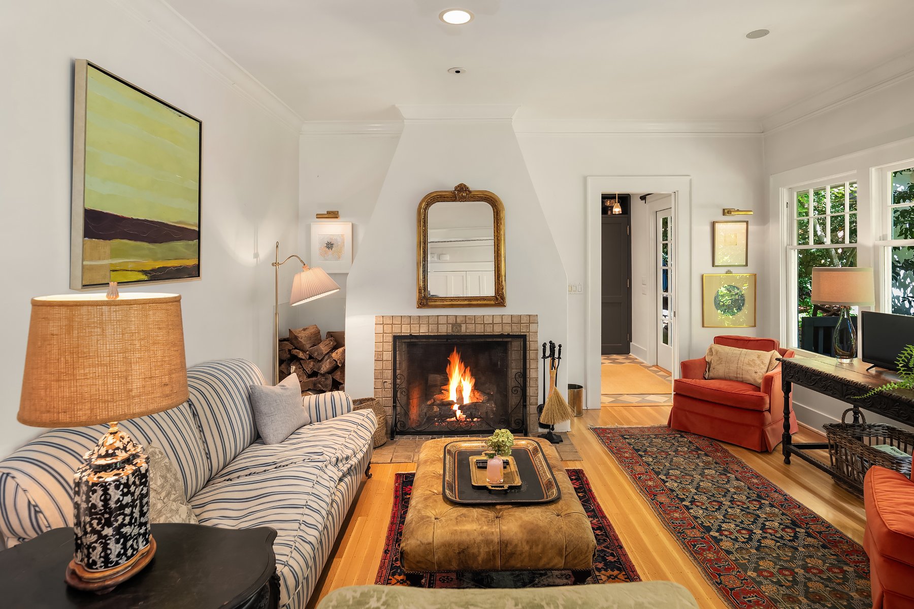 A cozy living room with a lit fireplace, striped sofa with pillows, a brass lamp, and a coffee table with decorations. There are artworks on the walls and large windows with trees outside, creating a warm, inviting atmosphere.