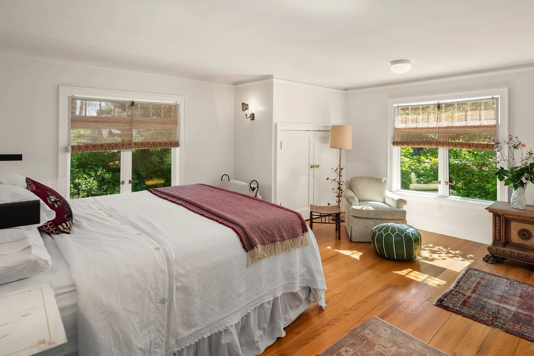 A bright bedroom with white walls, large windows with brown shades, a white bed with a red throw blanket, a beige armchair, a floor lamp, a small wooden table, green ottoman, and a floral vase on a wooden dresser, with sunlight streaming through the 