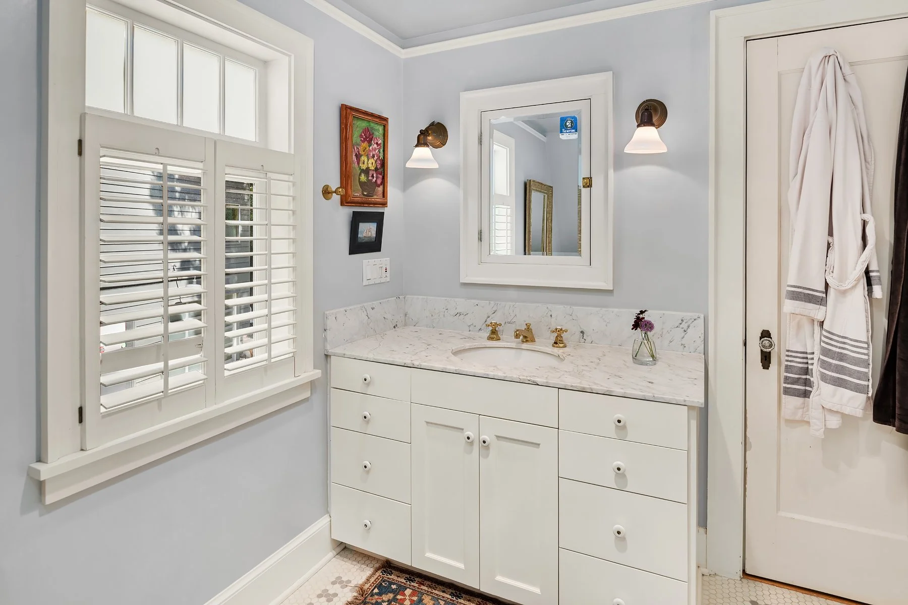 Bathroom with white vanity, marble countertop, gold fixtures, window with white shutters, mirror, wall lights, small vase with flowers, and a robe hanging on the door.