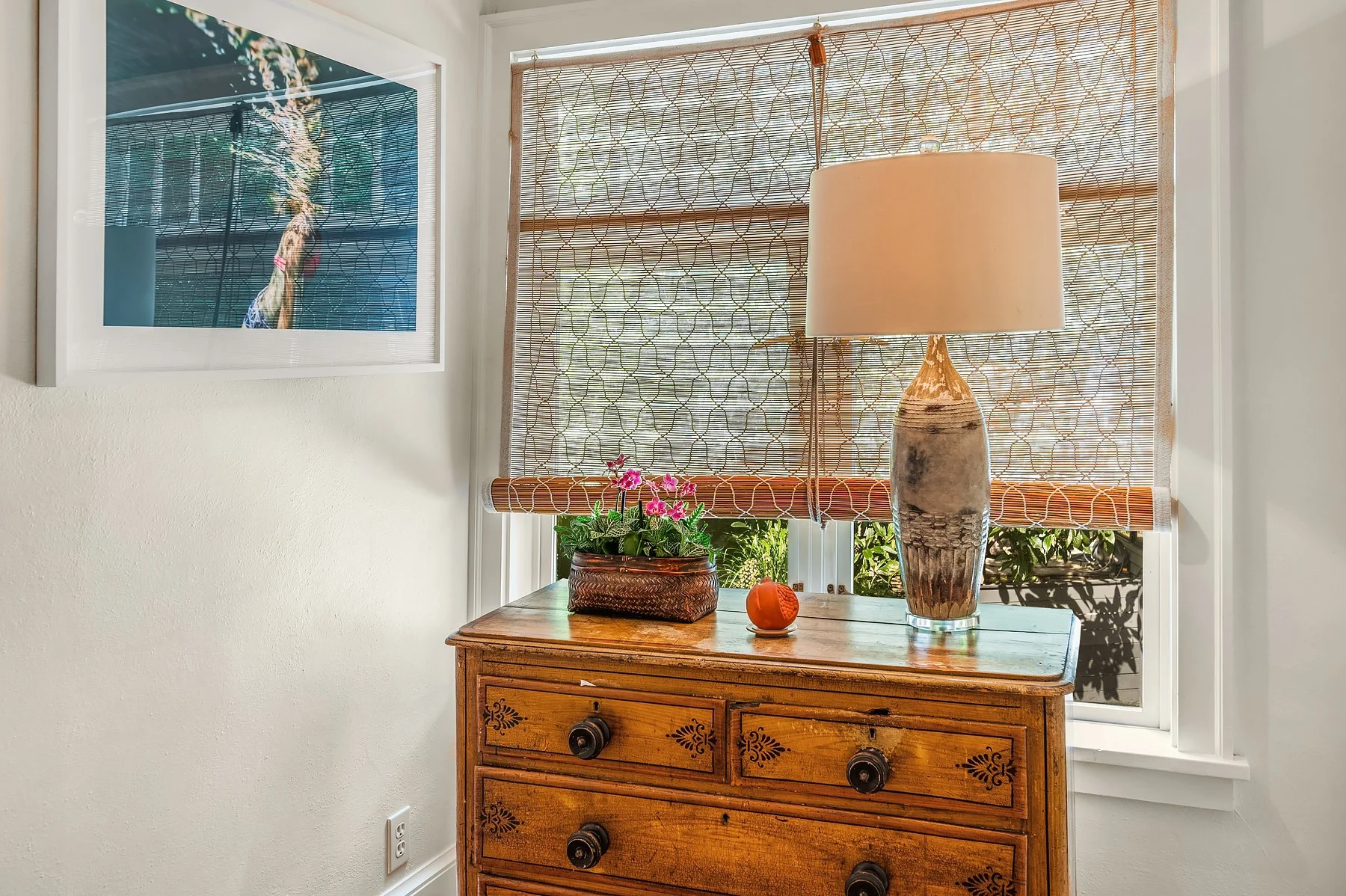 A wooden dresser with four drawers, a ceramic table lamp, a potted plant with pink flowers, and small decorative items. Behind it, a window with patterned bamboo blinds lets in sunlight, with greenery visible outside. A framed picture of a tiger is o