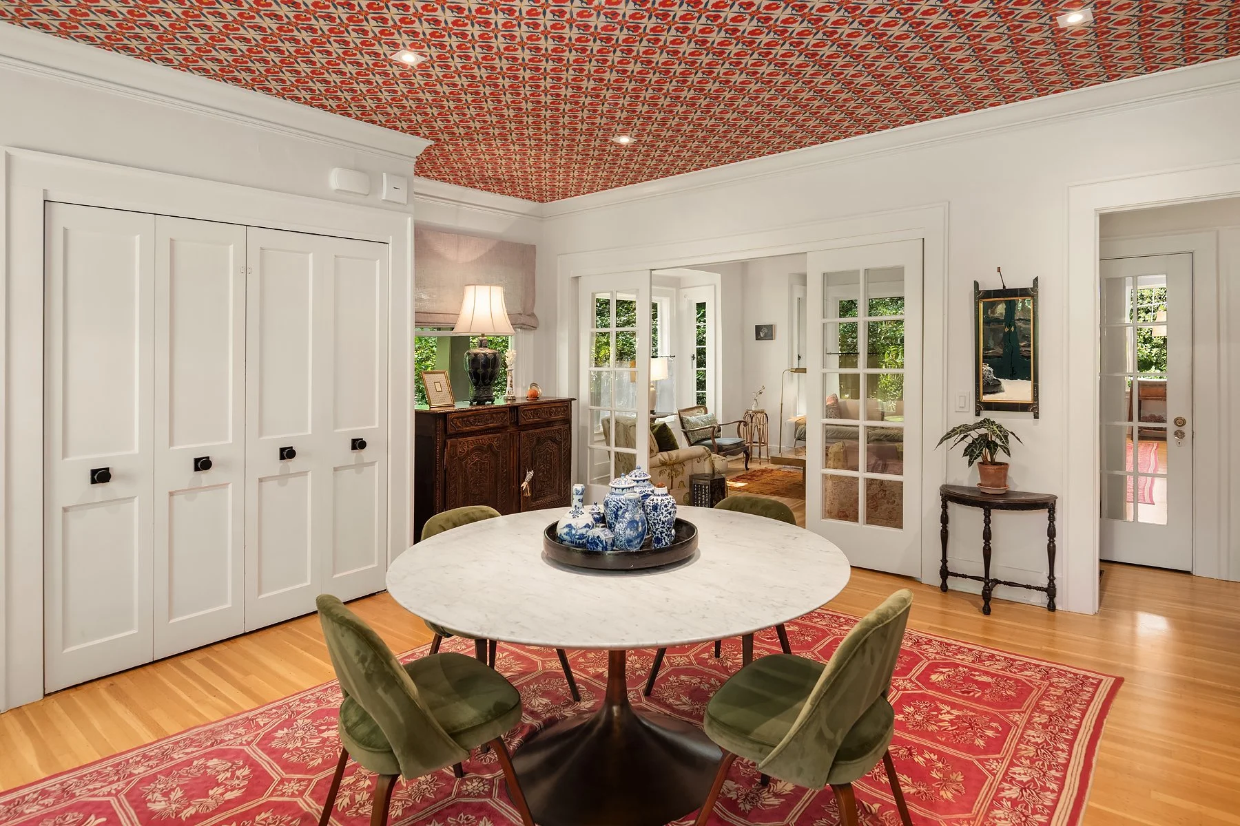 Dining room with a marble-topped round table surrounded by six green velvet chairs, a red patterned rug, and Chinese porcelain vases on a tray. The ceiling has a colorful patterned design, and there is a wooden sideboard, a table lamp, and multiple w