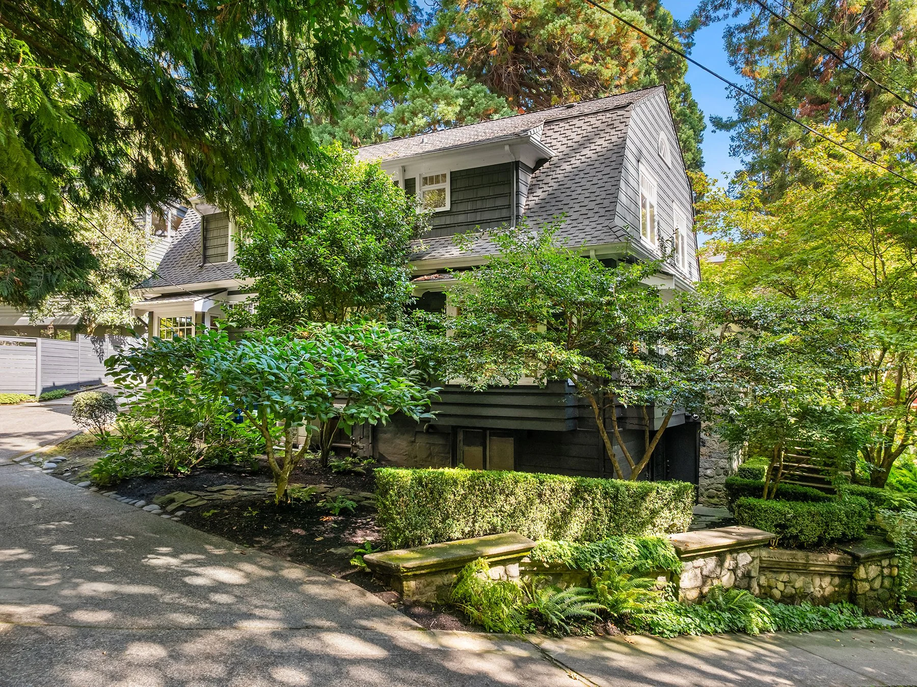A two-story house with dark gray siding and a steep shingled roof, surrounded by lush green trees and bushes, with a driveway in the foreground.