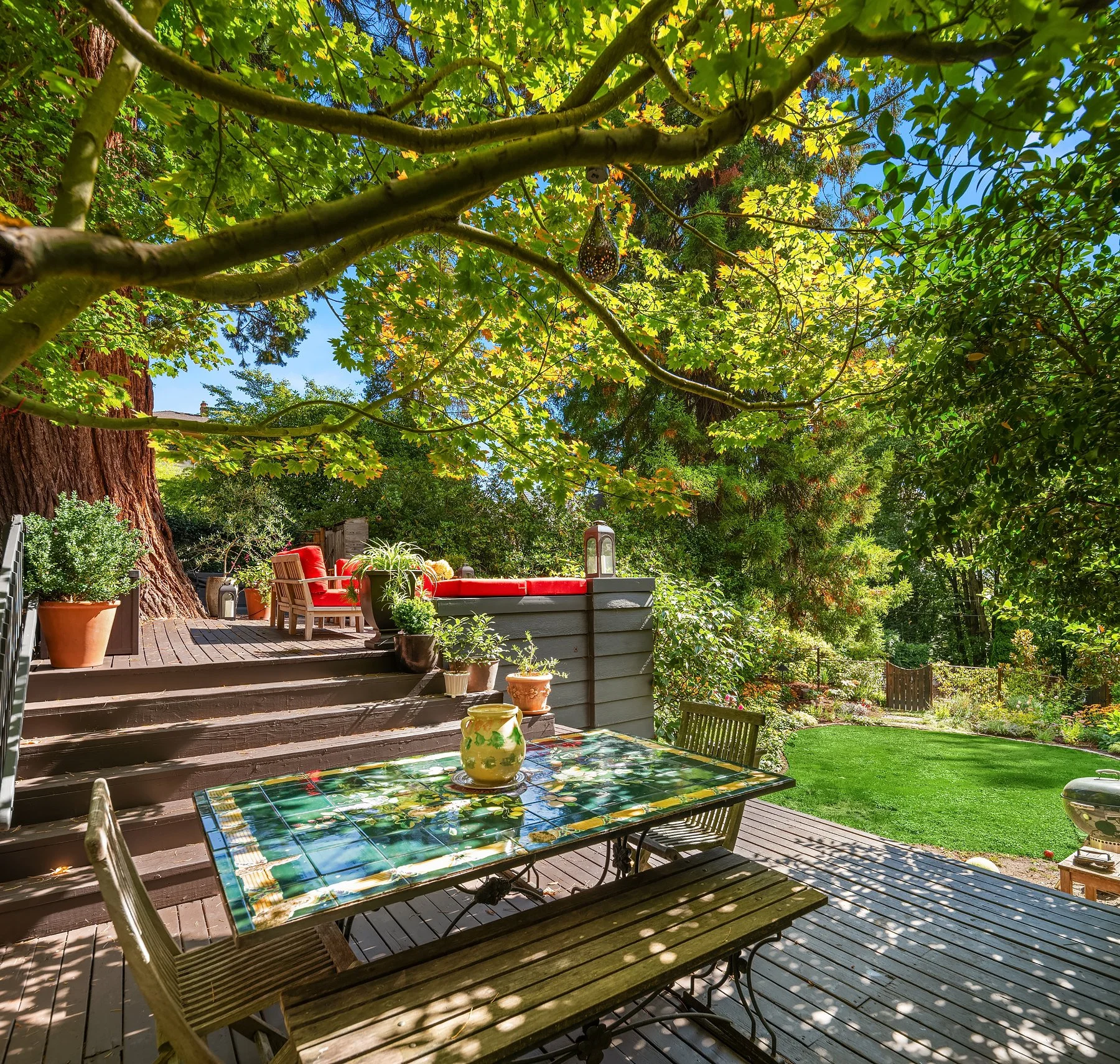 A backyard patio area with wooden steps leading to a garden with lush greenery and trees overhead. There is a glass-topped table with a yellow decorative vase, wooden chairs, potted plants, and a red cushioned seating area. The scene is sunny with a 