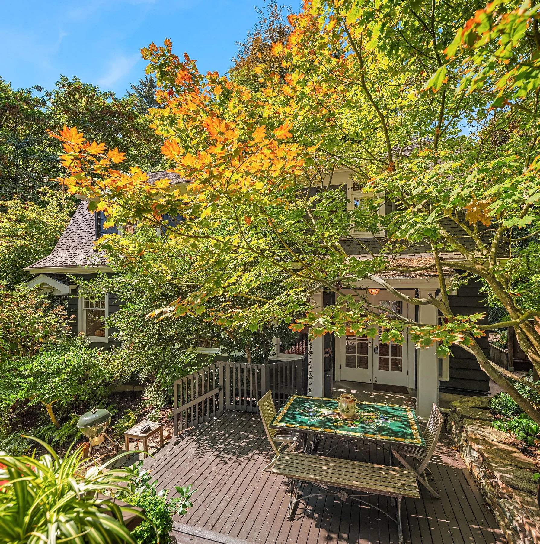 A backyard patio with a wooden deck, a table with a colorful floral tablecloth, and chairs, surrounded by lush green and some orange leaves from nearby trees. A house with gray siding and large windows is visible in the background, with a small fence