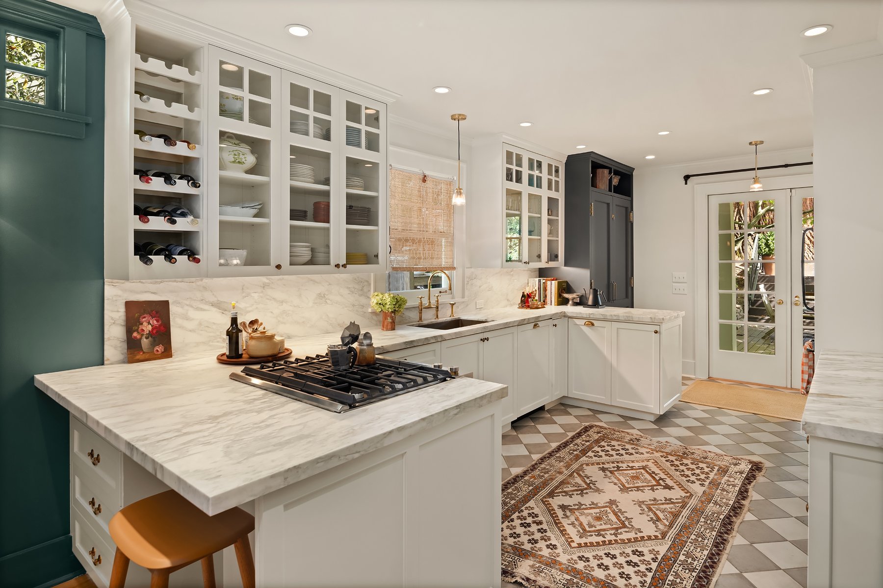 Bright kitchen with white marble countertops, white cabinets with glass doors, and a black cabinet. A door with glass panes leads outside. The kitchen has a patterned rug, a wooden stool, and various kitchen items like wine bottles, plants, and books