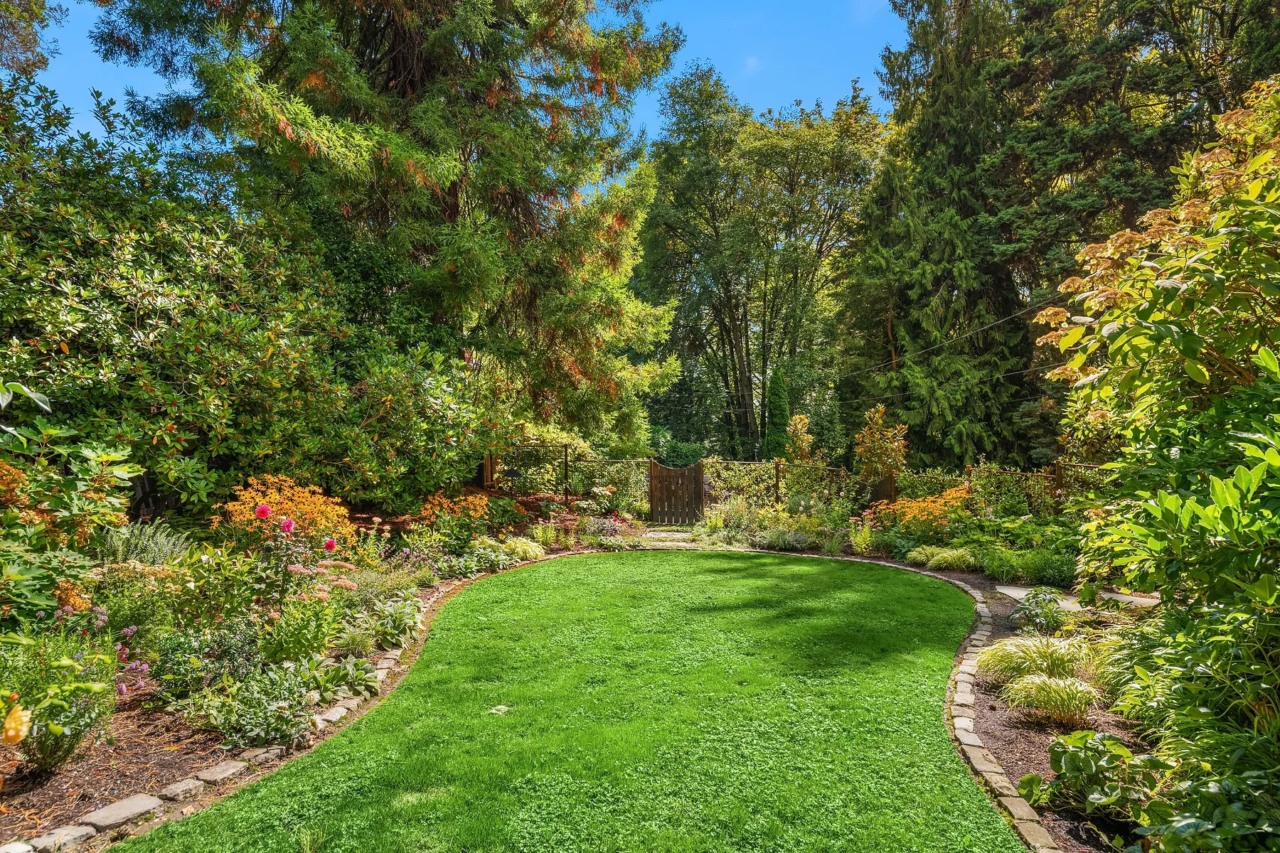 A manicured garden with a curved grassy lawn bordered by colorful flower beds and lush green trees under a clear blue sky.