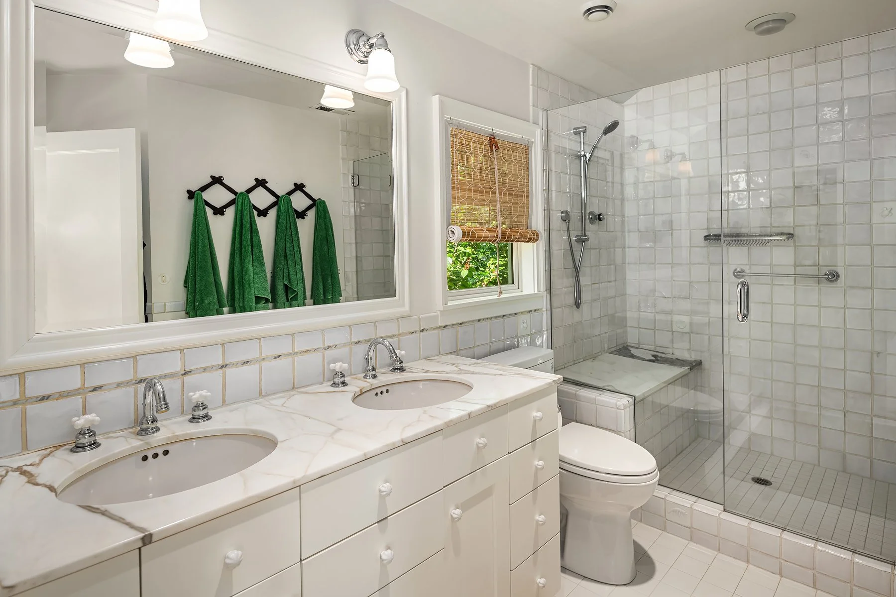 Bathroom with double sink vanity, large mirror, and glass-enclosed shower with tiled walls and a small window with bamboo shade.