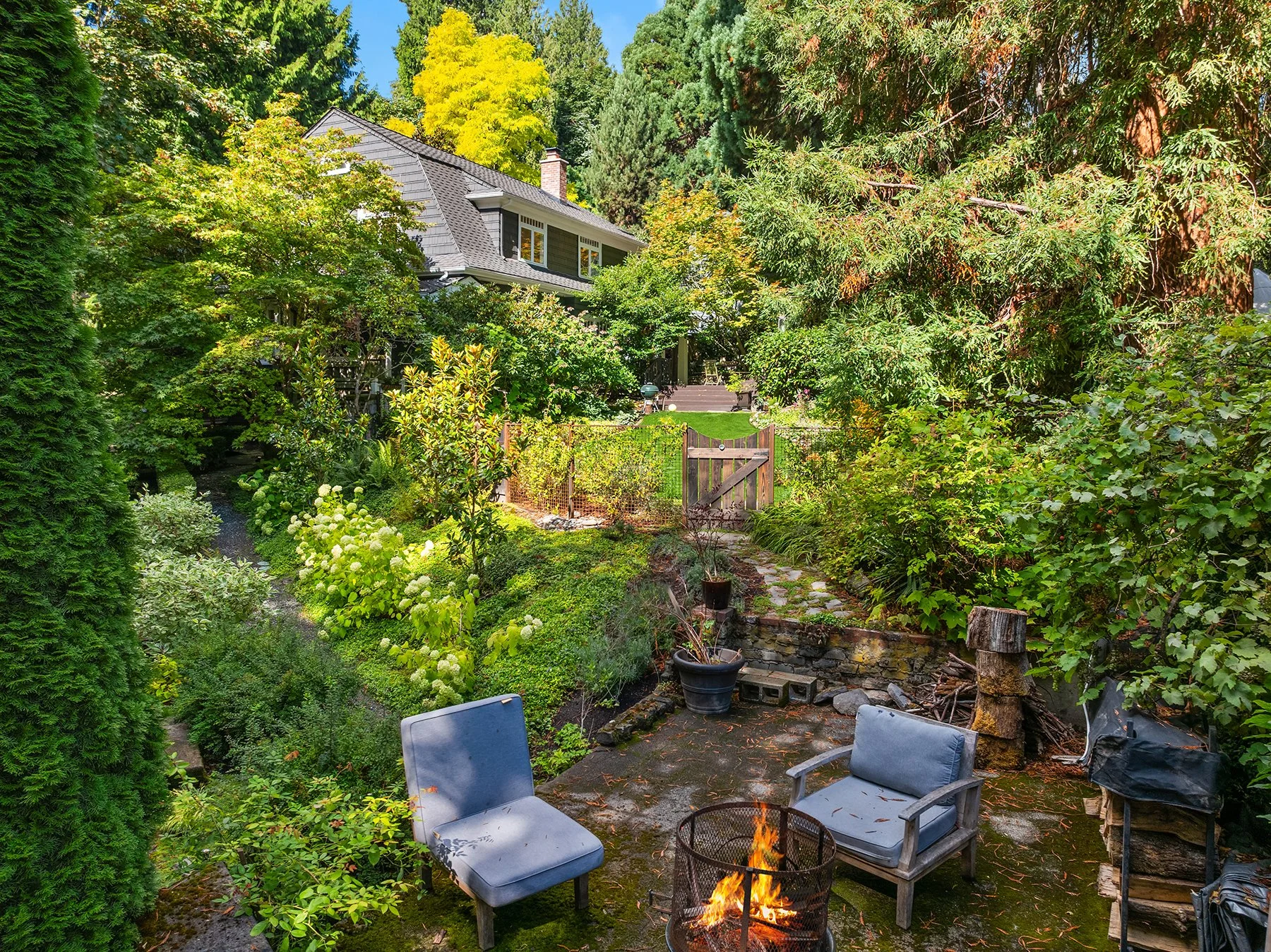 A backyard garden with lush green trees and plants, a stone pathway, and outdoor furniture including a small fire pit with a fire, chairs, and a wooden stump. There is a house partially visible in the background surrounded by tall trees and vibrant f