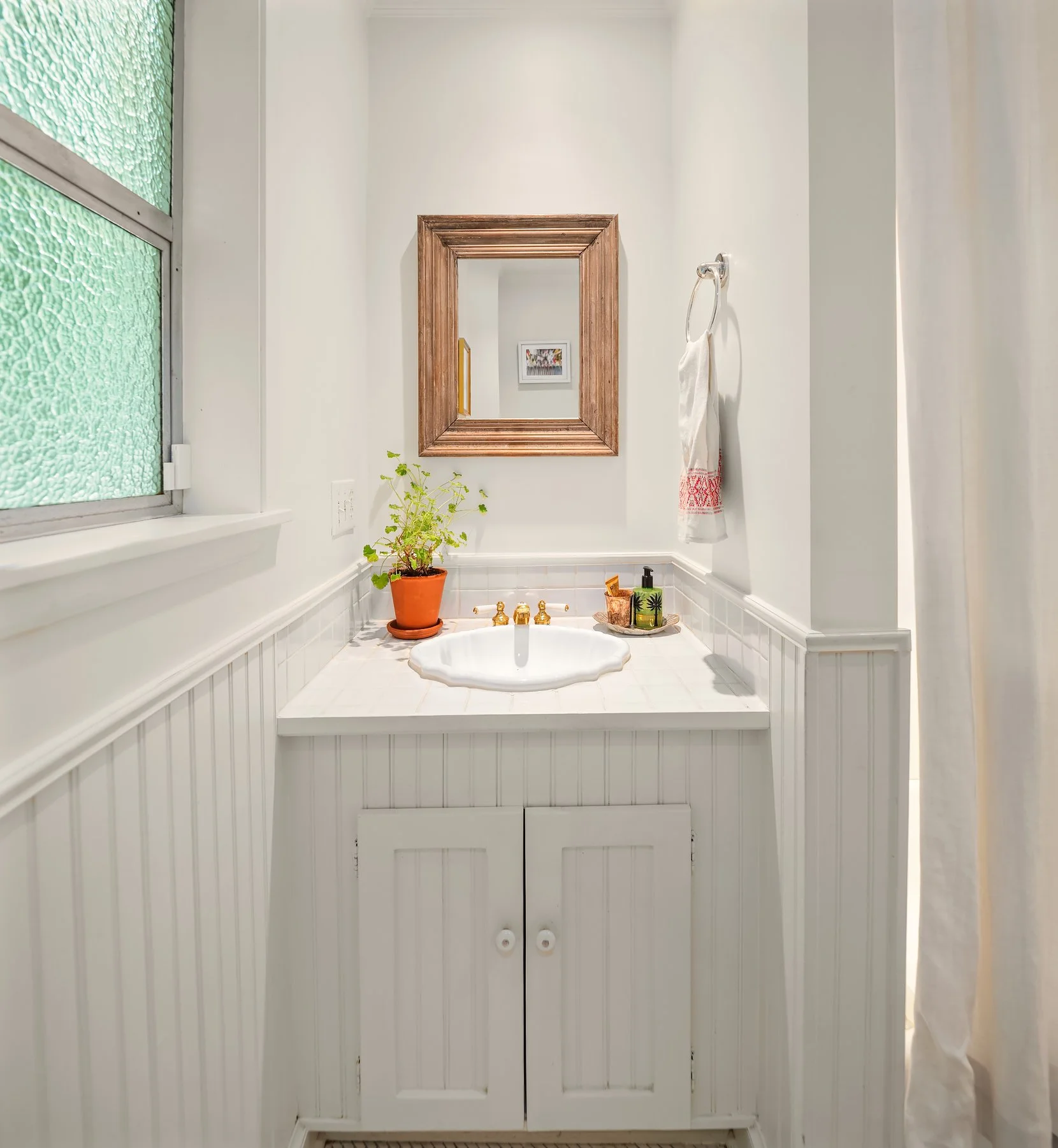Bathroom with a white vanity, a wooden-framed mirror, a window with textured glass, a potted plant, and toiletries on the counter.