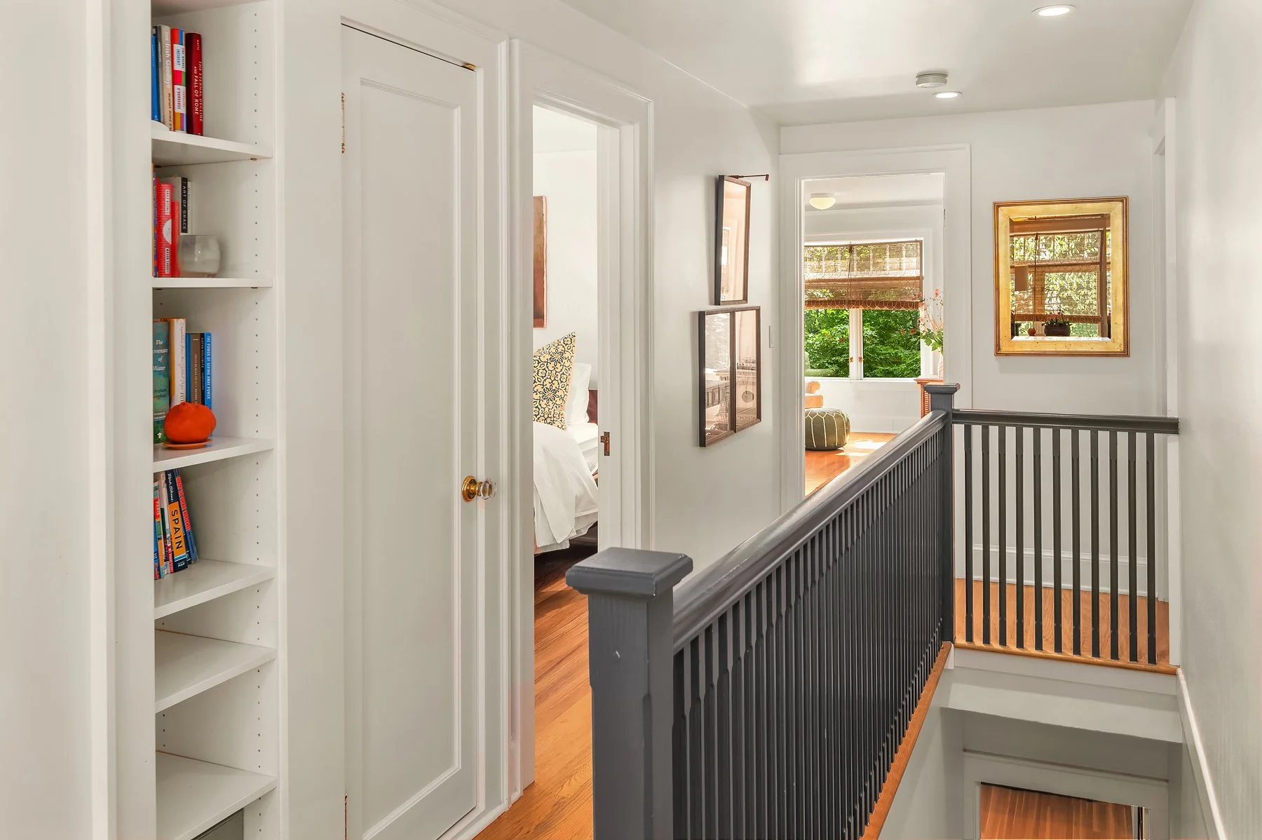 A hallway in a house with white walls, a hardwood floor, a dark gray wooden railing, and several framed pictures. Visible rooms include a bedroom with a bed and a sunlit sitting area with large windows and a round pouf.