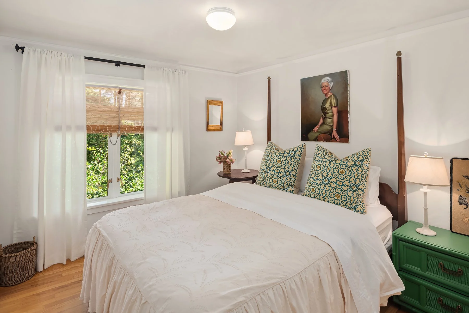 A bedroom with white walls, white curtains, and a large window overlooking greenery. There is a bed with white bedding and large decorative pillows, two bedside lamps, a small round wooden table with a lamp and flowers, and a painting of an elderly w