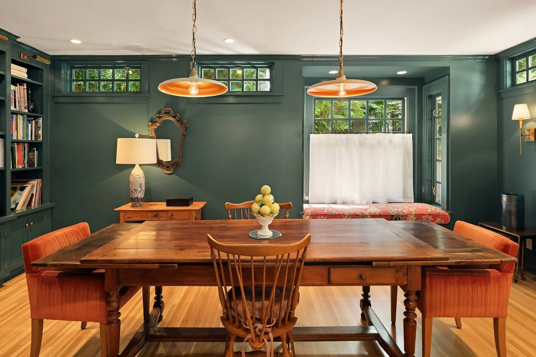 A dining room with a wooden table surrounded by eight chairs, including a captain's chair at the head. The table has a centerpiece of green and yellow apples in a white dish. The background features a wall with teal-green paint, a window seat with pi