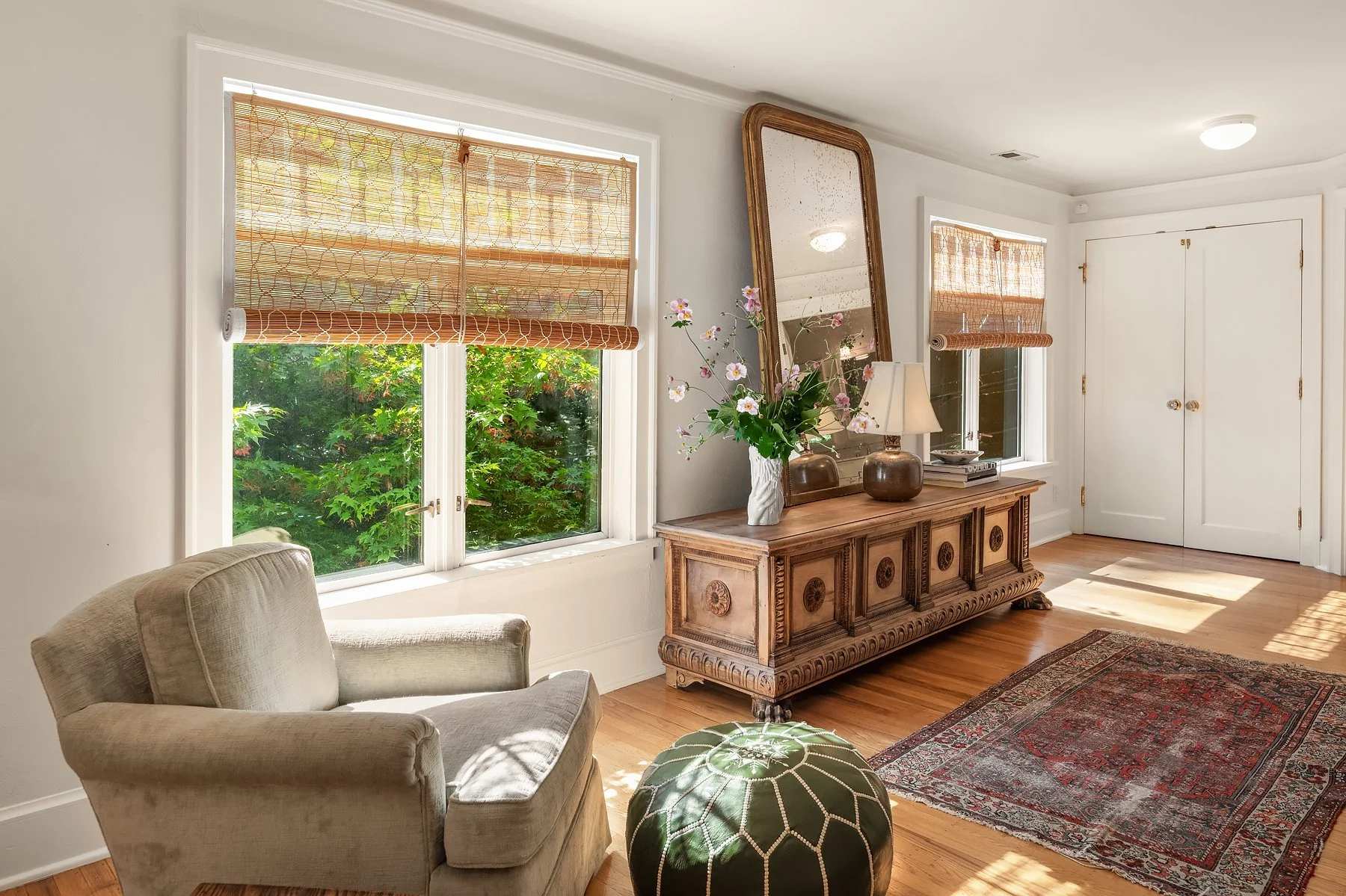 A cozy living room with two large windows covered with bamboo blinds, a beige armchair, a wooden sideboard with an ornate design, a large mirror, a table lamp, a vase with flowers, an area rug, and outdoor greenery visible through the windows.