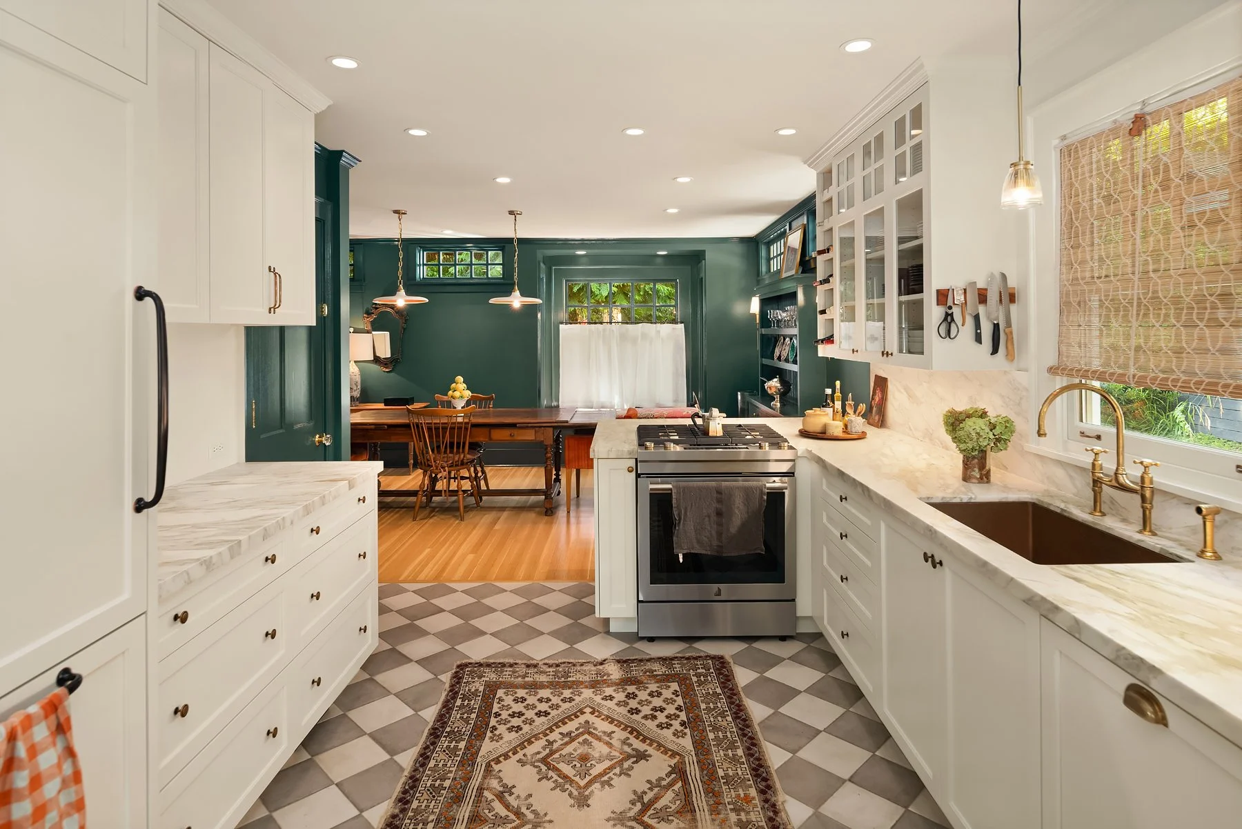 A bright, open kitchen with white cabinets, a marble countertop, a gold faucet, and a brown kitchen sink. The floor has black and white checkered tiles with a decorative area rug. In the background is a dining area with green walls, a wooden table, a