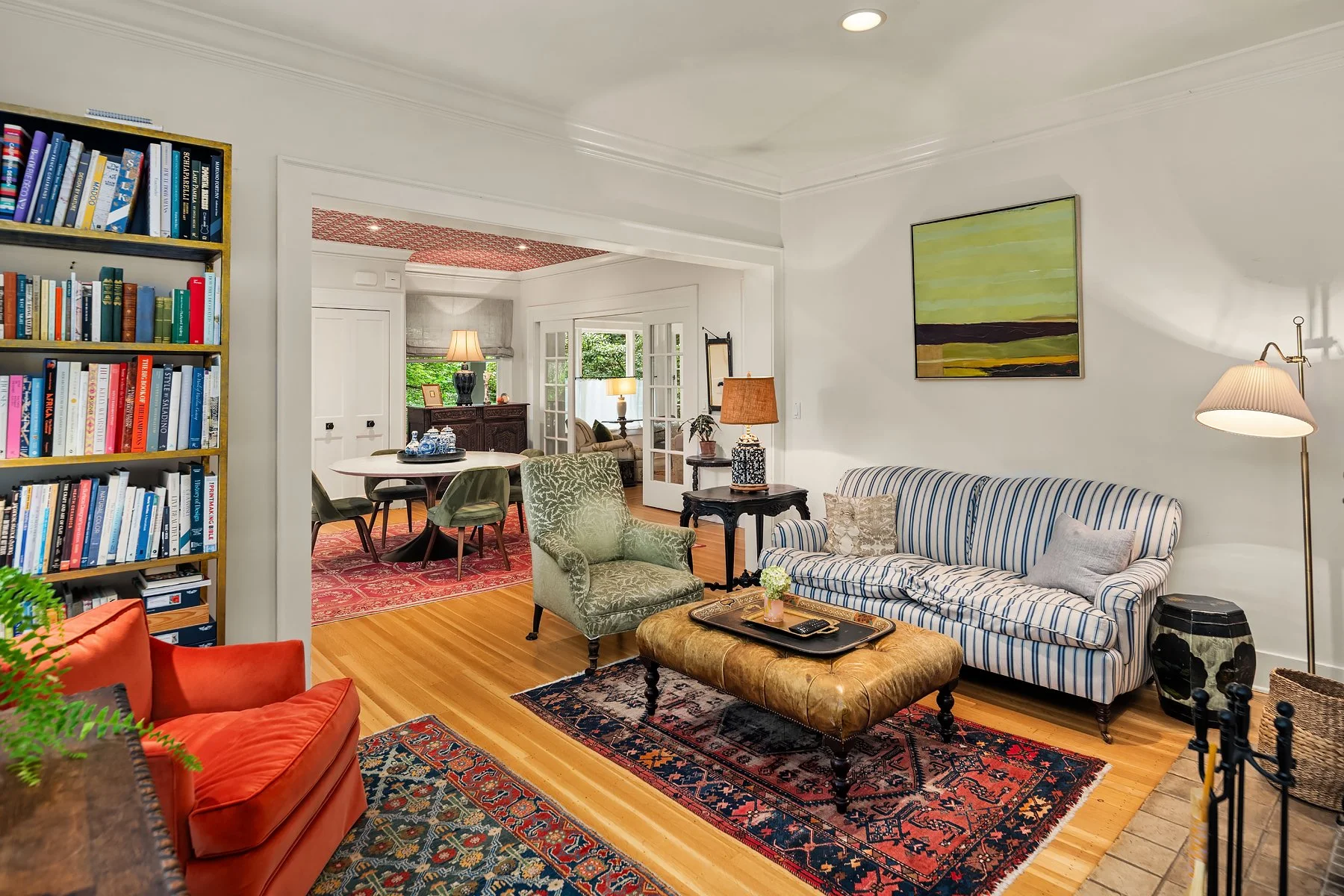 Living room with striped sofa, green armchair, side tables with lamps, and a bookshelf, leading to a dining area with a round table and chairs, hardwood floors, and artwork on the walls.