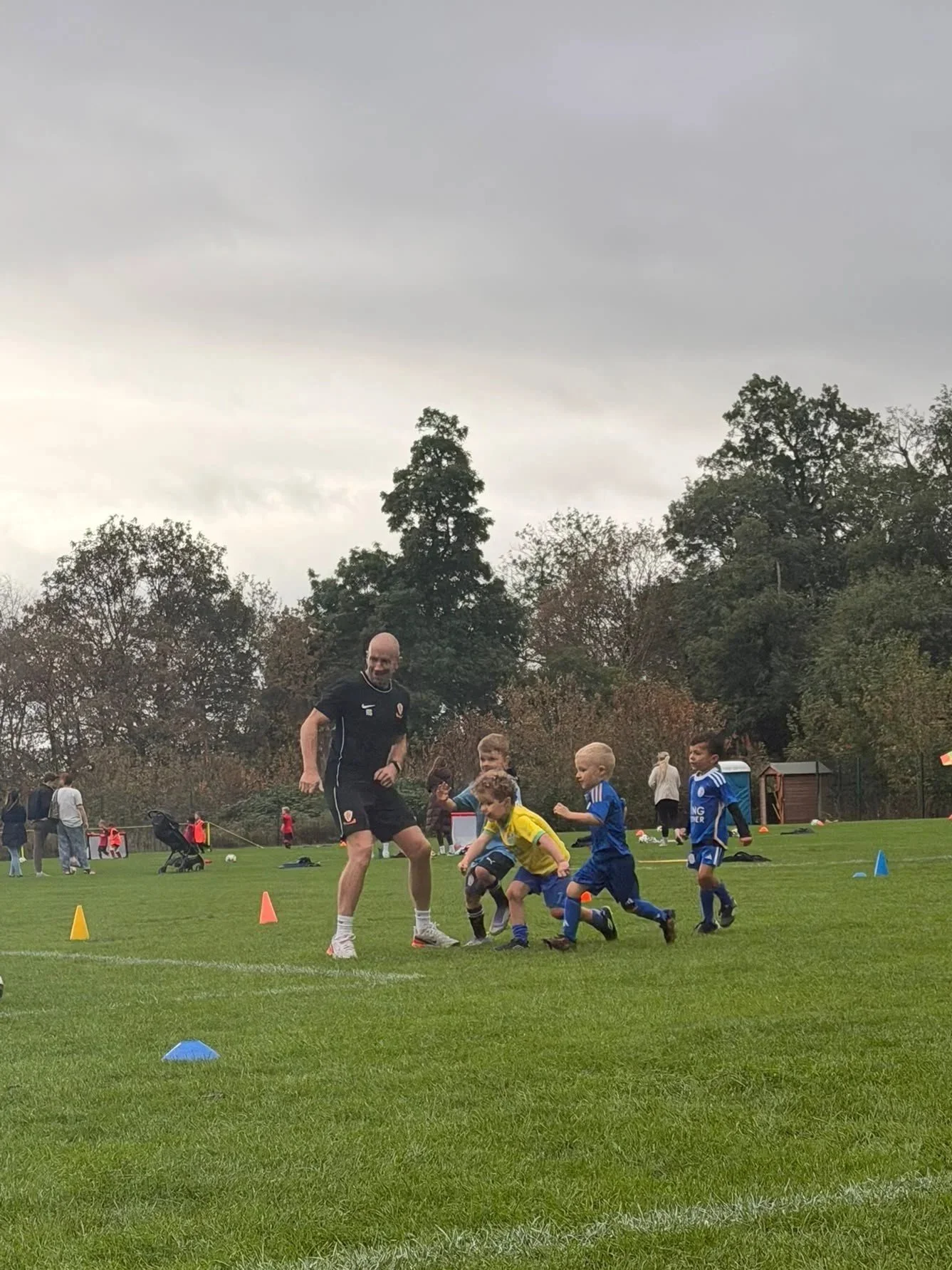 Children playing soccer on an outdoor field under a cloudy sky, with a coach or referee leading them. There are orange and blue cones on the grass and people in the background.