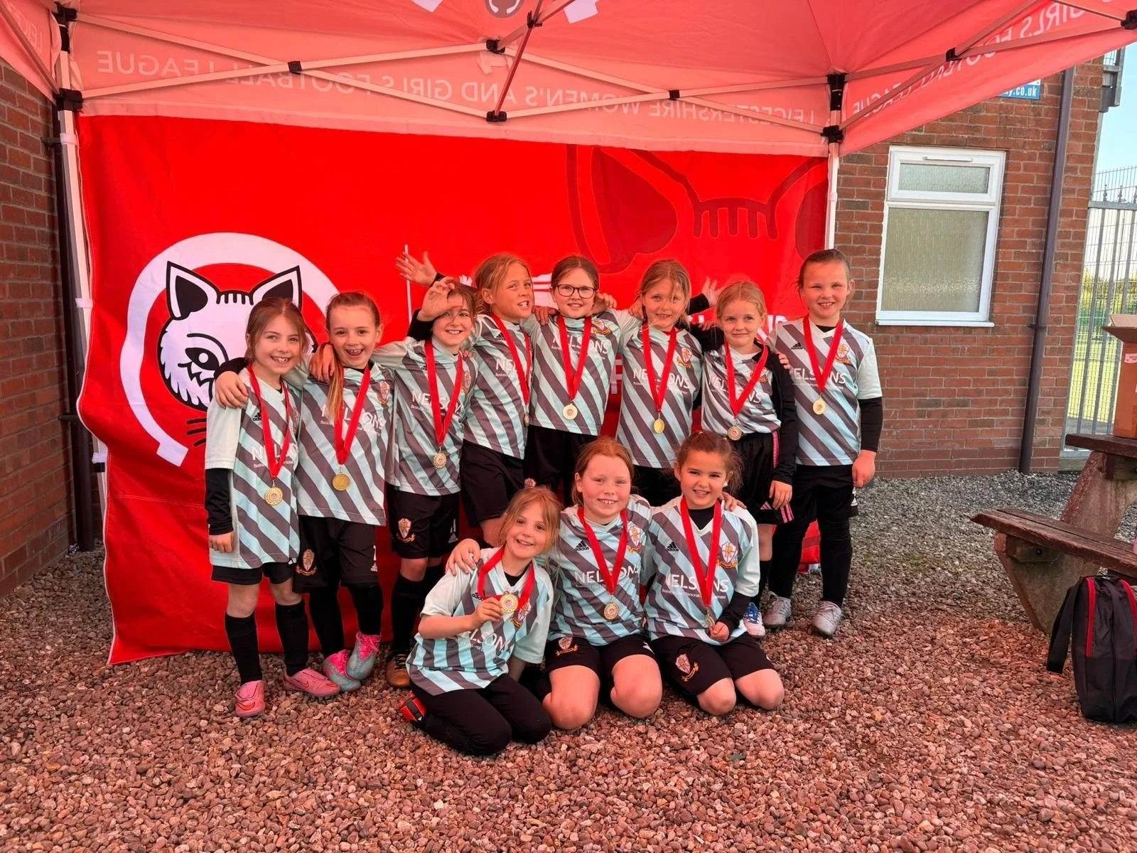 Group of young girls in sports uniforms with gold medals around their necks, posing for a photo under a red canopy with a lion logo in the background.