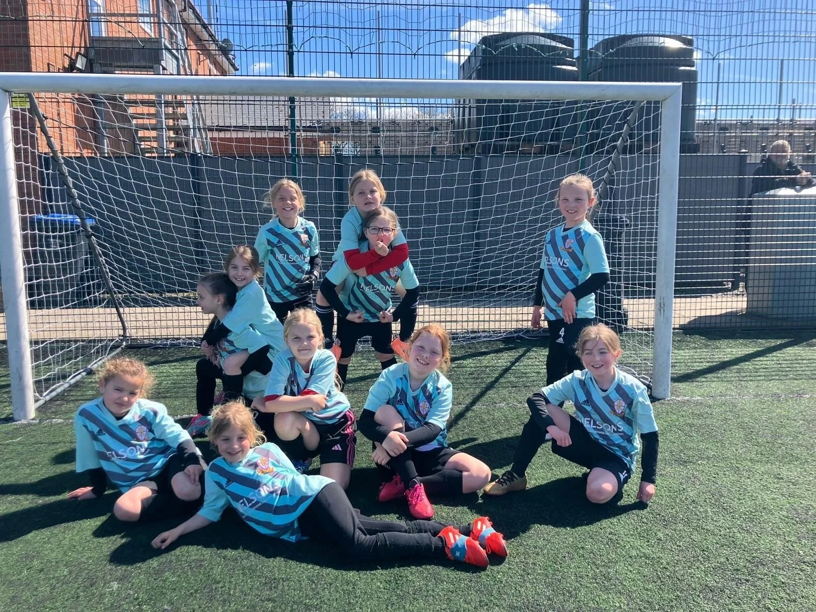 A group of young girls in soccer uniforms posing in front of a soccer goal on an artificial turf field on a sunny day.