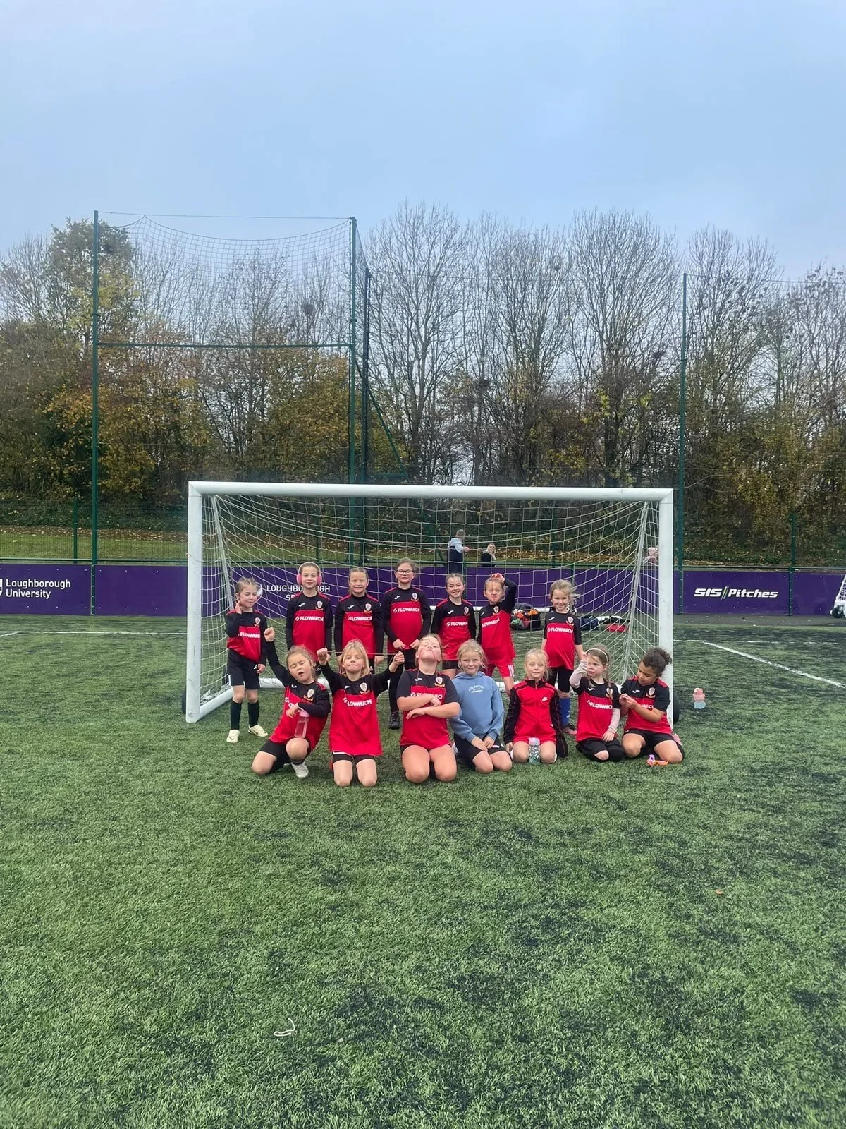 Group of young girls wearing red and black soccer uniforms posing for a team photo in front of a soccer goal on a green field. Some girls are standing, others are kneeling or sitting.